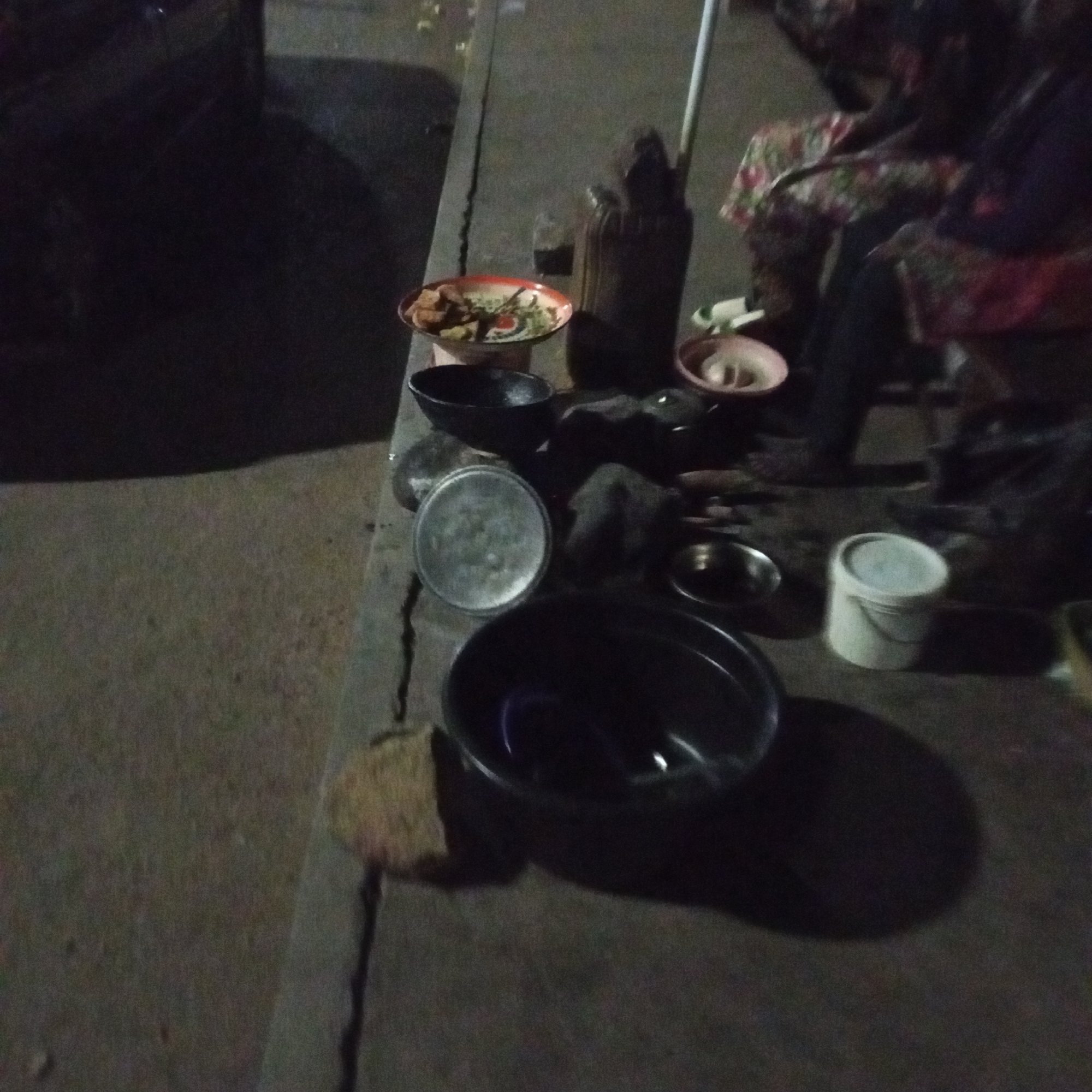 The image depicts an outdoor scene, likely at night, in what appears to be Bukuru, Nigeria, based on the implied context. Several containers are placed on a concrete surface that resembles a sidewalk. A large black bowl dominates the foreground, while a covered metal container, several smaller bowls, and a white bucket are also visible. A seated person is partly visible on the right side of the frame. In the background, there is a container full of food. The overall scene suggests a temporary food preparation or selling setup on a sidewalk.