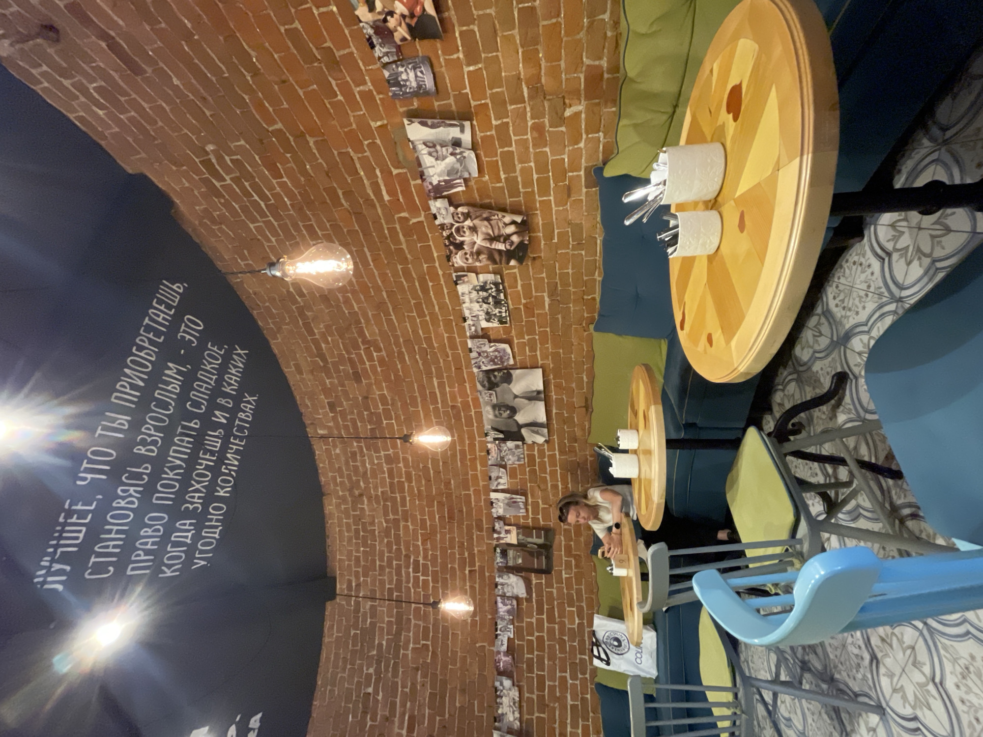 This image shows the interior of a cafe in Krasnoyarsk, Russia. The cafe has a circular layout with exposed brick walls and a dark ceiling adorned with white text and hanging light bulbs. The text on the ceiling is in Russian and translates to "The best thing you get when you become an adult is the right to buy sweets whenever you want, in any quantity." The cafe is furnished with plush blue and green seating, round wooden tables, and chairs of various colors, including blue and gray. Black and white photographs are displayed on a line running along the brick wall. A young woman is seated at a table, looking at her phone. The floor is tiled with a grey and white patterned design.