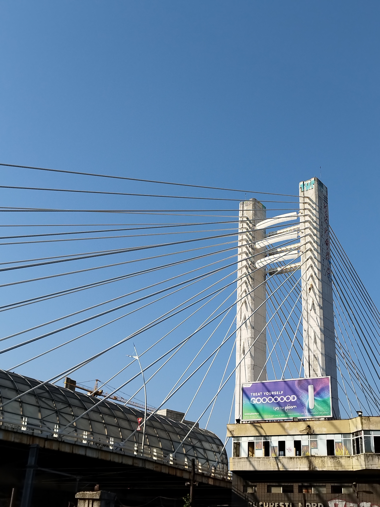 This is a low-angle, outdoor, daytime photograph of the Basarab Bridge in Bucharest, Romania, under a clear blue sky. The focus is on the bridge's massive concrete pylon and its numerous suspension cables. A billboard advertising "GOOOOOD LyO for BLOOM" is prominently displayed on the side of a building attached to the bridge structure, which also features graffiti. To the left, part of the bridge's elevated roadway, covered with a translucent material, is visible, along with a construction crane in the background. The lower part of the image shows the underbelly of the bridge and some urban decay with further graffiti. The overall scene depicts a mix of modern infrastructure and elements of urban life.