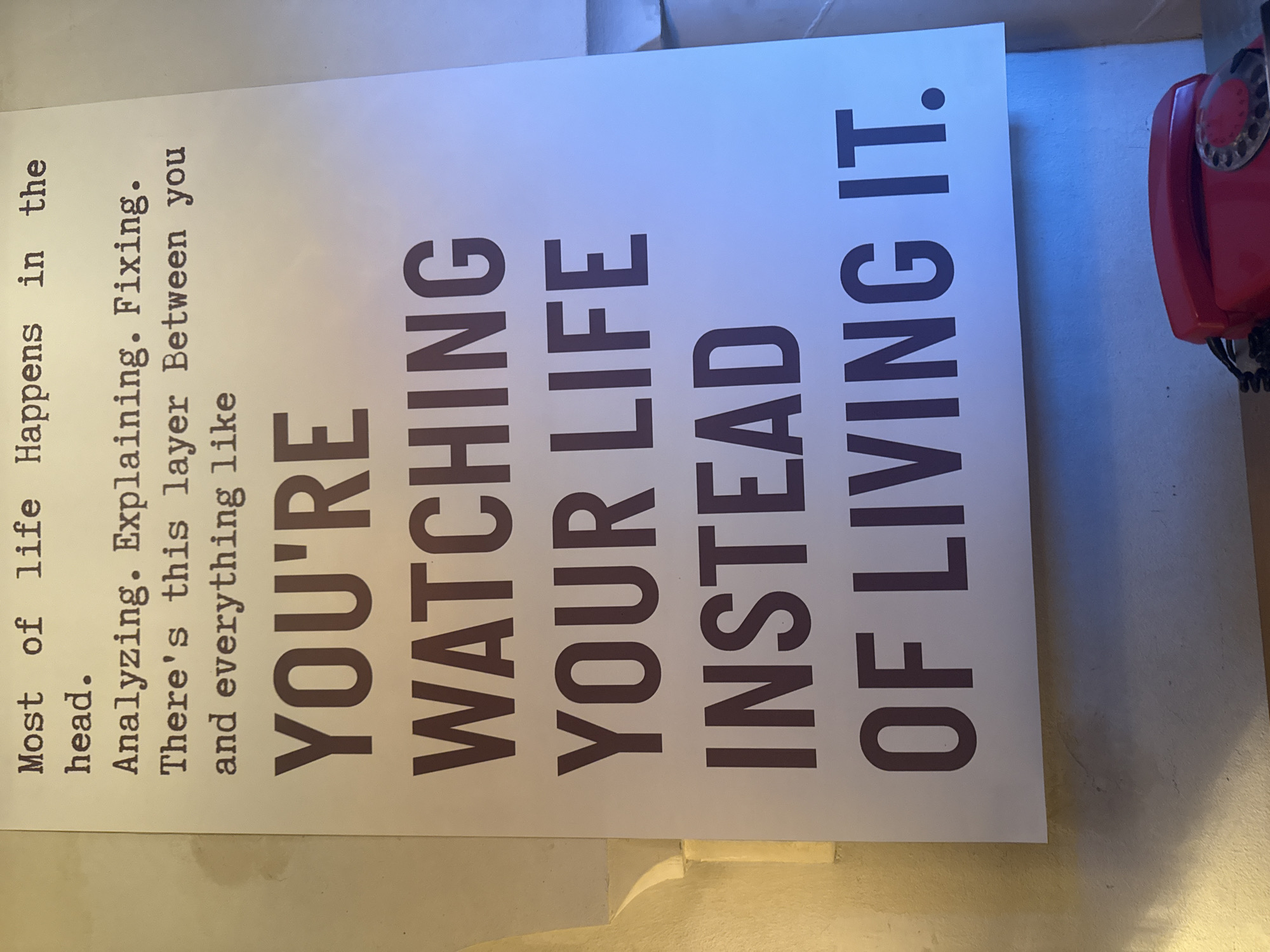 A vertically oriented poster with typography is prominently displayed in a brightly lit environment. The poster features a motivational message in a serif typeface: "Most of life Happens in the head. Analyzing. Explaining. Fixing. There's this layer Between you and everything like YOU'RE WATCHING YOUR LIFE INSTEAD OF LIVING IT." The poster is predominately white with dark text, suggesting it's mounted on a wall or similar surface.
To the right, a partially visible red rotary dial telephone is present, giving context to the setting. The lighting seems diffuse, evenly illuminating the scene. Based on the artifacts, it is possible that the picture was taken in the city of Yerevan, Armenia.