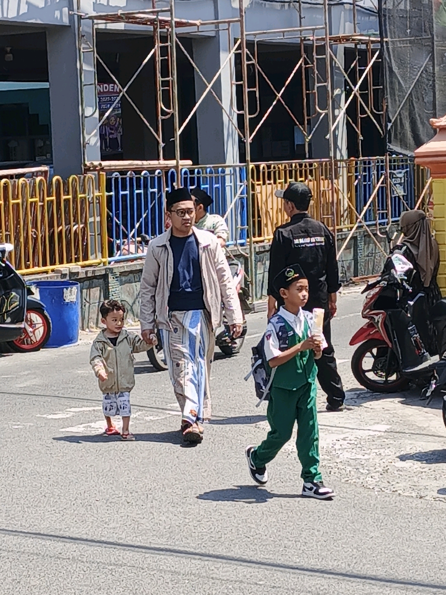 In Ketegan, Indonesia, on a sunny day, a group of people are walking on a street. In the foreground, a man in a light-colored jacket and patterned pants, wearing glasses and a black skullcap, walks hand-in-hand with a young boy. The boy is wearing a light-colored hooded jacket and patterned pants. To their right, another young boy, wearing a white shirt, green vest, green pants, and a black cap with a logo, walks forward holding a light-colored object. Behind them, two individuals are visible near motorcycles. One is a person wearing a black jacket with writing on the back, and the other is a woman in a brown headscarf. In the background, scaffolding and construction netting suggest ongoing building work, with a fence in front of the structure. Several motorcycles are parked or moving in the scene. The street is paved, and there is a blue cylindrical bin on the left side of the road.