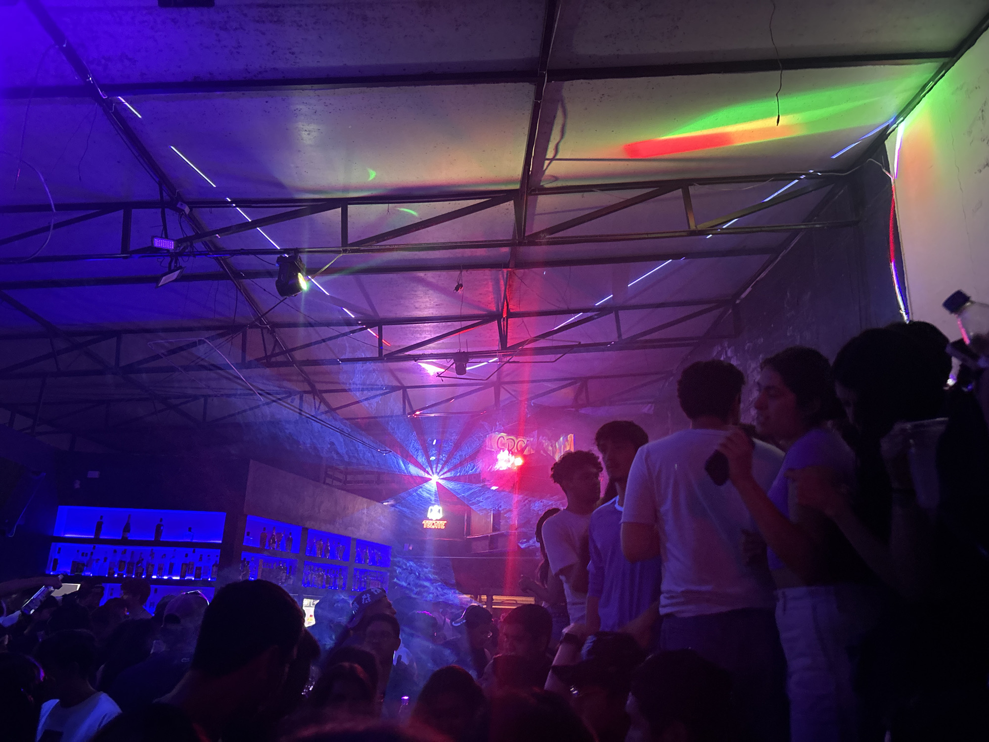 The image depicts a crowded and lively indoor club or bar scene at night. Numerous people are visible throughout the space, mostly in the mid-ground and foreground, mingling and interacting. The venue is illuminated by a vibrant array of artificial lights, casting strong purple, blue, red, and green hues across the room and ceiling. Laser lights cut through the air, creating distinct lines and dynamic patterns, including a prominent starburst design on the left side, suggesting the presence of atmospheric smoke or haze.
On the left, a long bar area is brightly lit with a glowing blue light, showcasing multiple shelves stocked with bottles of alcohol. The ceiling features an industrial-style metal truss structure with suspended lighting fixtures, including spotlights and rectangular LED panels. The ceiling panels reflect the colorful lights intensely, particularly red and green streaks in the upper right corner.
A sign displaying the text "TECATE" is clearly visible on the left side, positioned above the bar area. The overall atmosphere is energetic and festive, typical of a busy nightlife establishment. The scene conveys a sense of enjoyment and social gathering taking place in an entertainment venue located in San Andrés Cholula, Mexico.