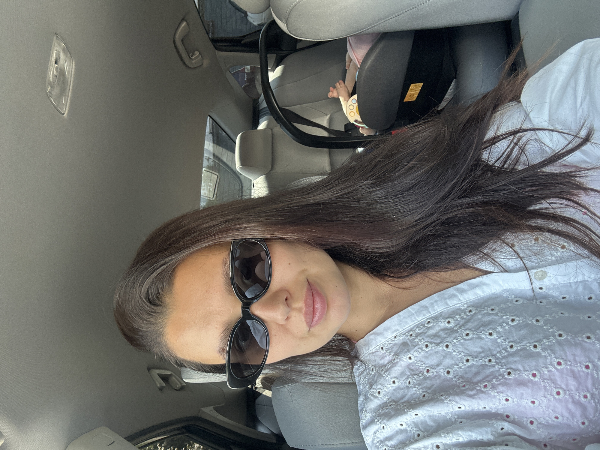 A woman with long, dark hair and black sunglasses is seated in the front passenger seat of a car, facing the camera. She is wearing a white eyelet shirt. In the backseat behind her, a baby is visible in a car seat, wearing a pink outfit. The interior of the car appears to be a standard sedan with gray upholstery. The scene is lit by natural sunlight filtering through the car windows. There is no discernible location in the city of Chaikovsky, Russia.