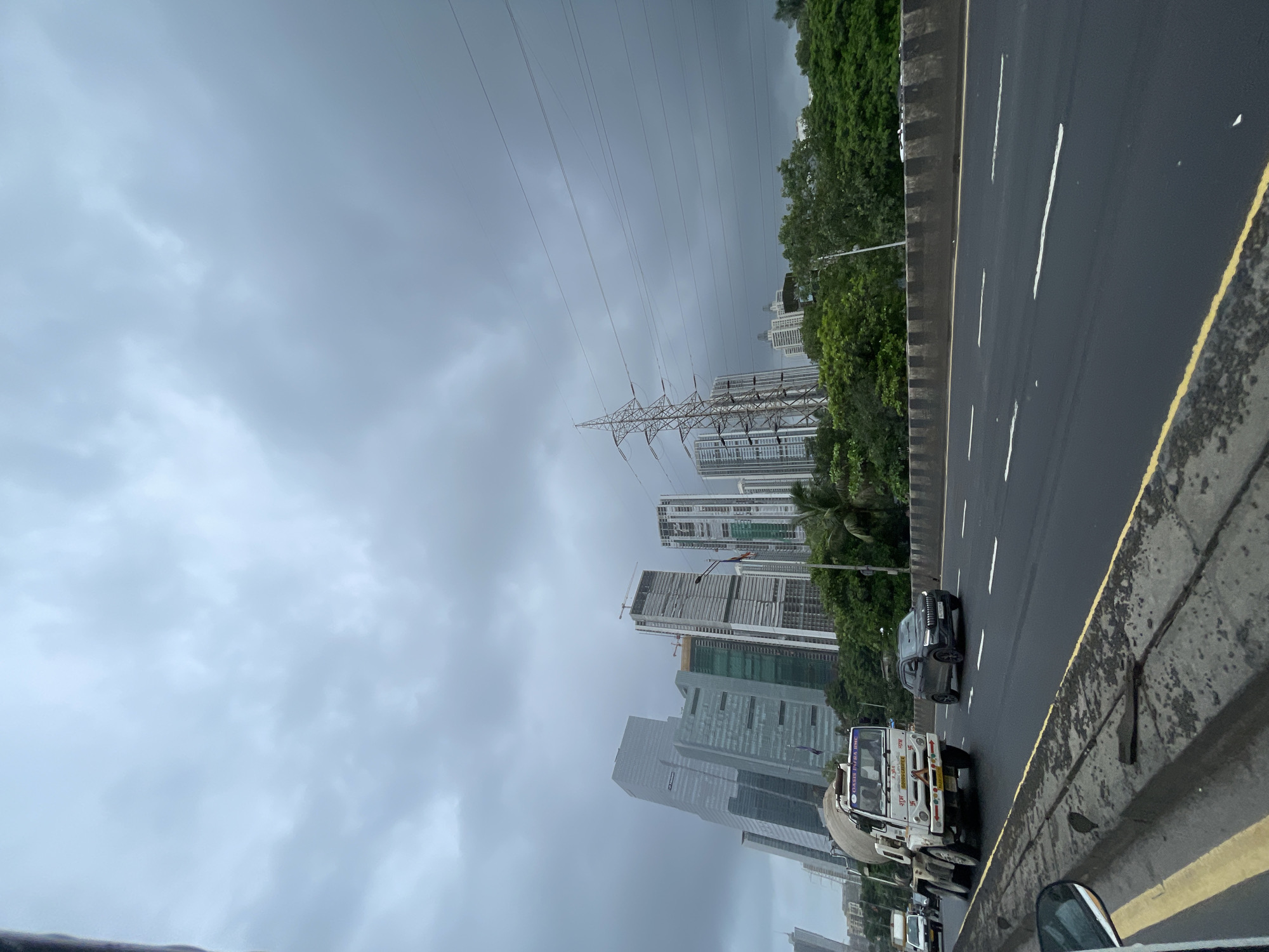 This is a vertical, eye-level, medium shot photograph taken from a vehicle on a highway in Mumbai, India, on a cloudy day. The highway is paved and has clear white lane markings and a yellow line bordering the right side of the road. Several vehicles are visible on the highway, including a white cement mixer truck in the foreground on the left, a dark SUV in the middle lane on the right, and a few other cars in the distance.
The background features a cluster of modern, tall skyscrapers, some of which appear to be under construction. Green trees and vegetation line the side of the highway, separating it from the buildings. A large power pylon with multiple wires strung between it and other structures is visible to the right of the skyscrapers. The sky is overcast with heavy, dark gray clouds. The overall impression is an urban highway scene in Mumbai, likely during monsoon season given the cloudy sky.
