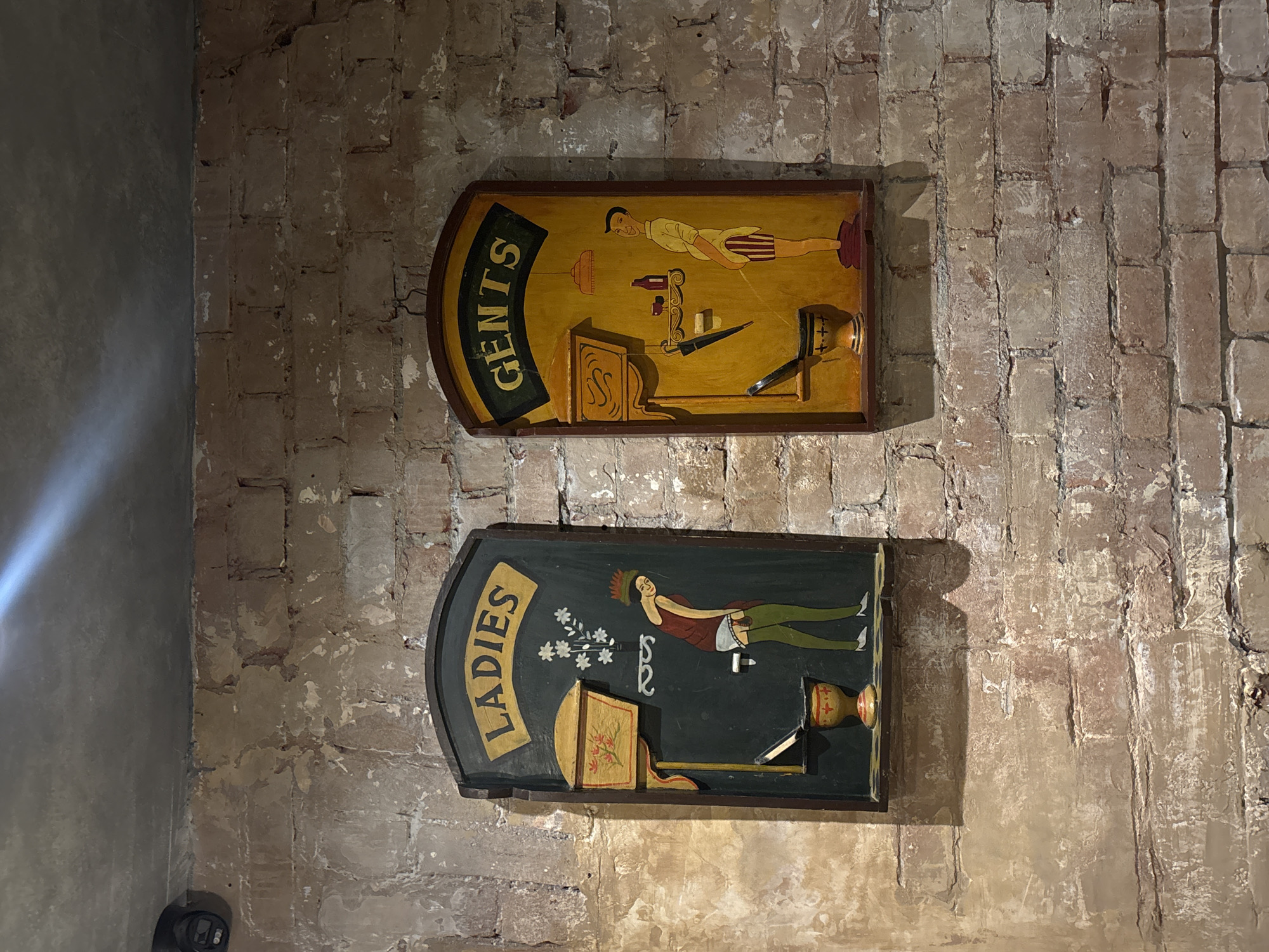Two wooden signs are mounted on a brick wall. The upper sign reads "GENTS" and features an illustration of a man and objects associated with a men's restroom, such as a shaving kit. The lower sign reads "LADIES" and features an illustration of a woman and objects associated with a women's restroom. The setting seems to be in Moscow, Russia, based on the style of the signs and the brick wall. The image shows restroom signs in a building with brick walls.