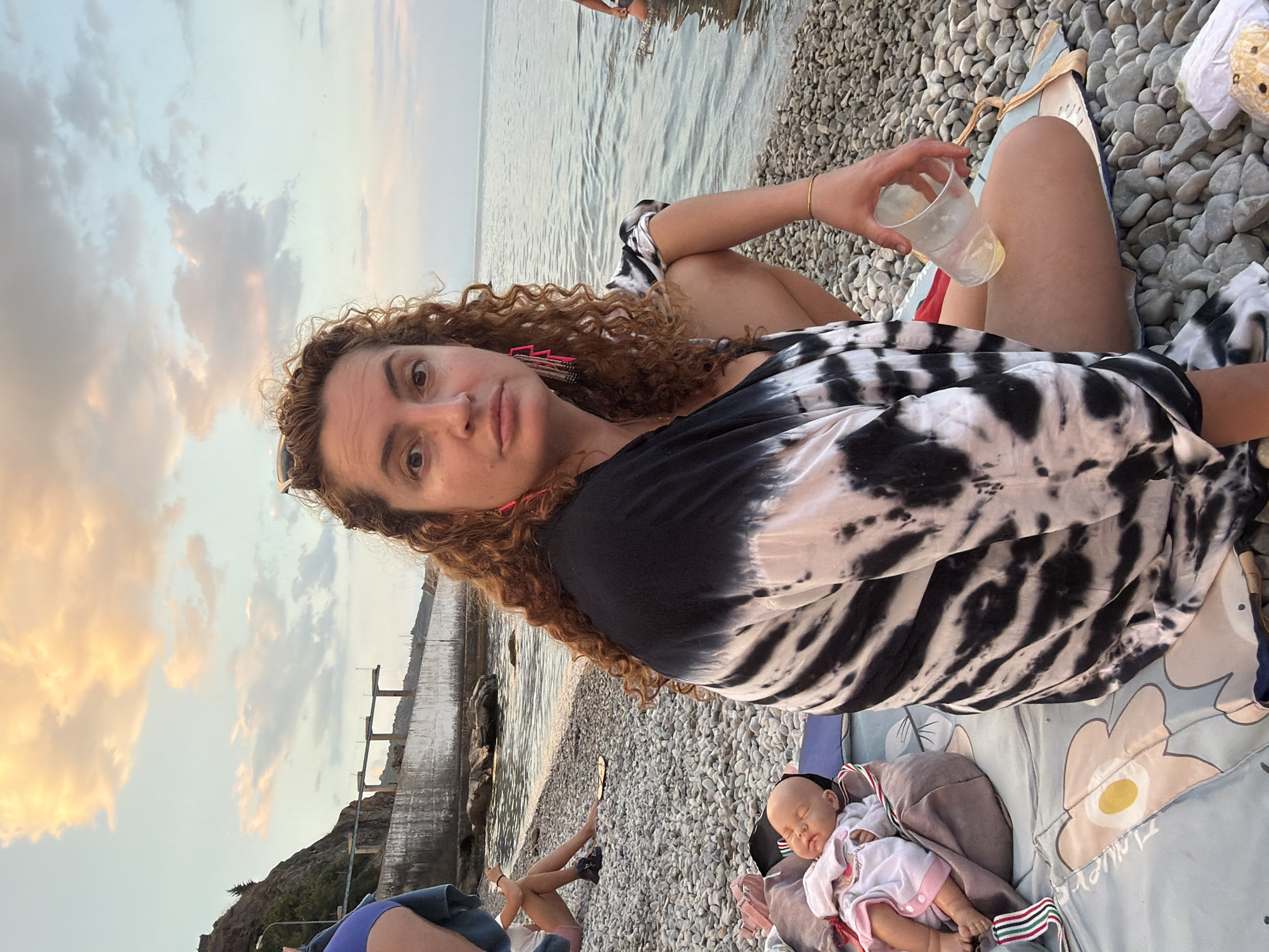 A woman with curly, auburn hair, wearing a black and white tie-dye top and bright pink earrings, sits on a pebble beach. She is holding a clear plastic cup with a yellowish liquid in her right hand. Next to her, on a patterned blanket, lies a baby doll dressed in a white outfit with pink trim. In the background, the sea meets the shore, and a concrete structure, possibly a seawall or promenade, runs along the coastline. The sky above is partly cloudy with hues of orange and pink, suggesting either sunrise or sunset. The location is identified as the city of Partenit, Ukraine. Other individuals are visible in the distance on the beach and in the water.