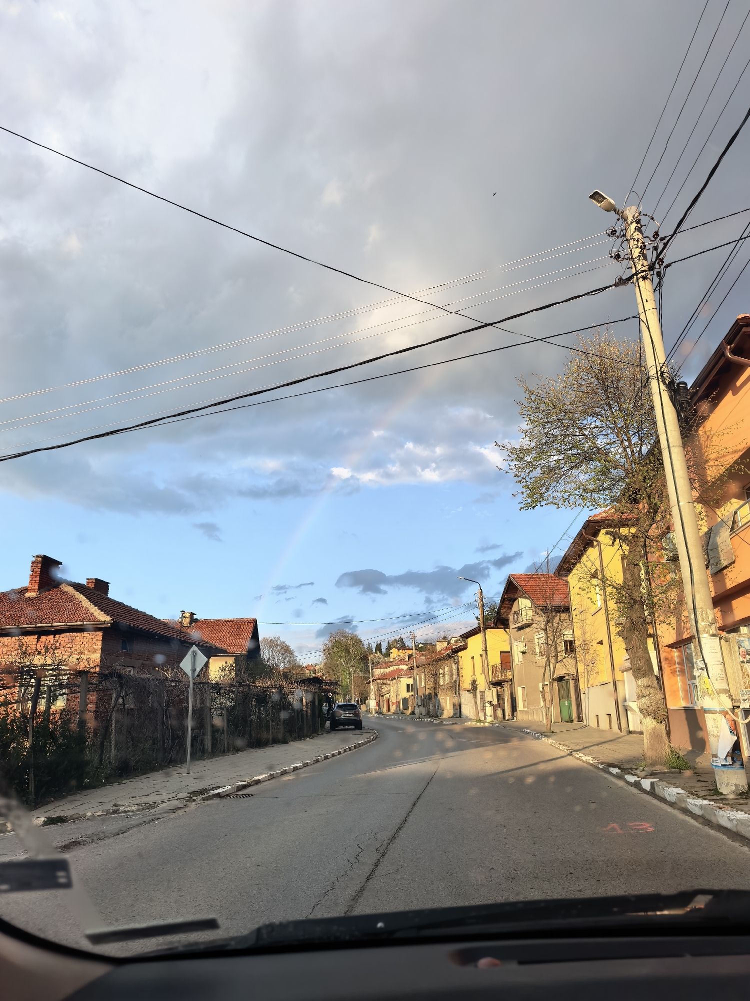 The image shows a view from inside a vehicle, looking down a narrow street lined with buildings in Unknown, Unknown. The buildings are a mix of styles and colors, including brick and stucco, with red tile roofs. A partial rainbow is visible in the sky between the buildings. Overhead, power lines cross the street, and a utility pole stands on the right side. The street is paved and appears to be somewhat worn. The sky is partly cloudy with patches of blue. A car is parked further down the street.