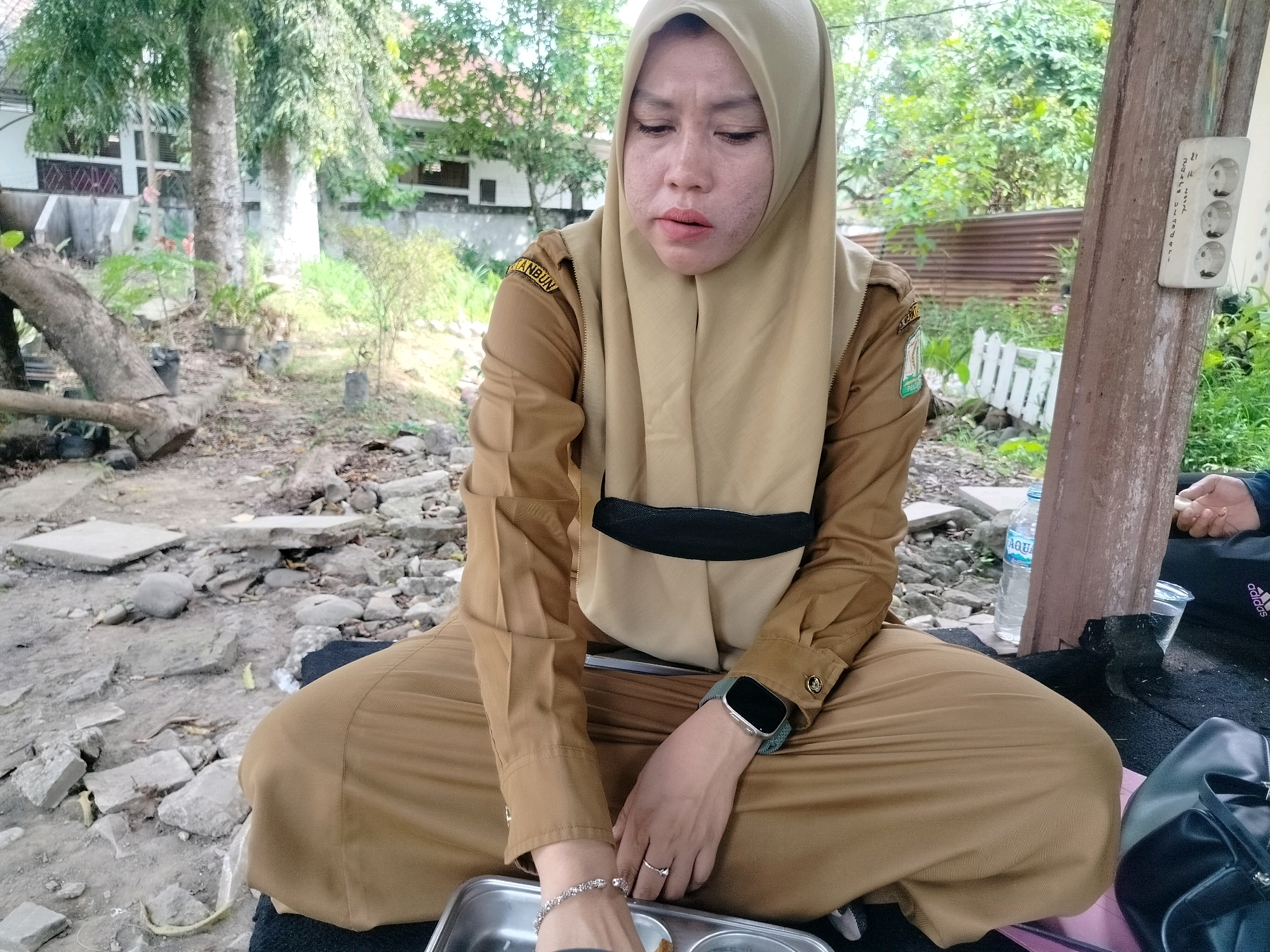 A medium shot captures a woman wearing a tan uniform with a hijab, sitting cross-legged on the ground outdoors in what appears to be a somewhat neglected area. She is looking down with her eyes closed, her lips slightly parted. Her left hand rests on her lap, adorned with a bracelet and a ring. Her right hand is also resting on her lap. She wears a smartwatch on her left wrist. Behind her, a wooden post with a three-socket electrical outlet is visible, with handwritten notes on the post. The environment is characterized by rough ground, scattered rocks, concrete debris, and some greenery. In the background, trees and a building are partially visible. A plastic water bottle and a clear plastic cup are on the ground near the woman, along with a black bag. The scene is set in Purwodadi, Indonesia.