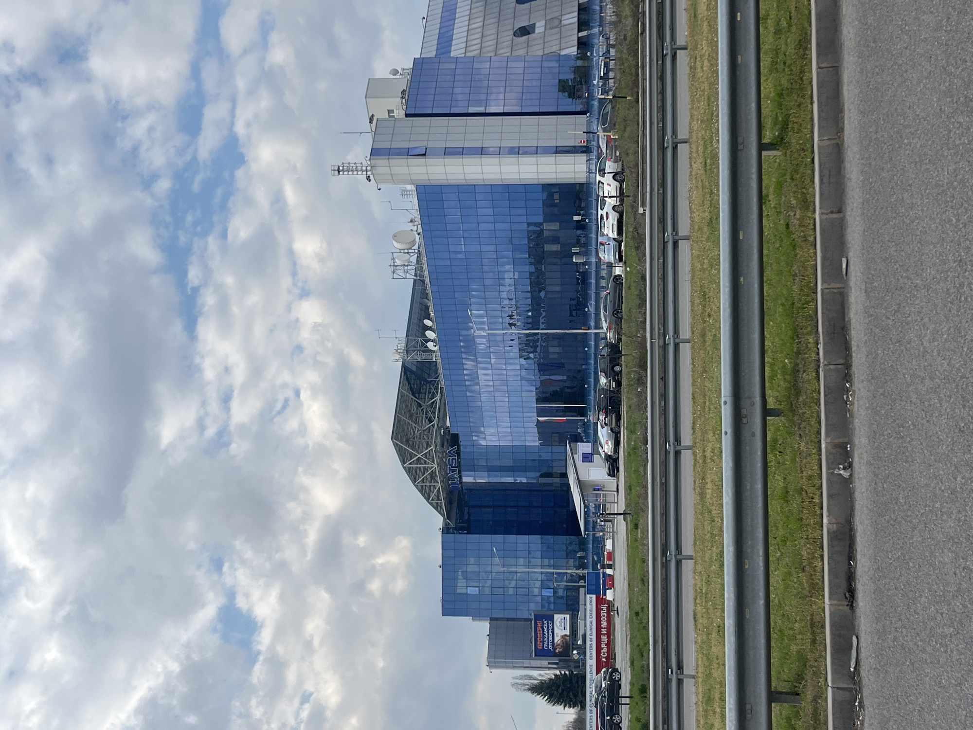The image depicts a tall, modern, blue-glass building complex situated in Sofia, Bulgaria. The building has satellite dishes and communication equipment on its roof. A highway runs alongside the complex, with a metal guardrail separating the road from a grassy area. Cars are parked parallel to the building. A billboard is visible near the building. The sky is partly cloudy.