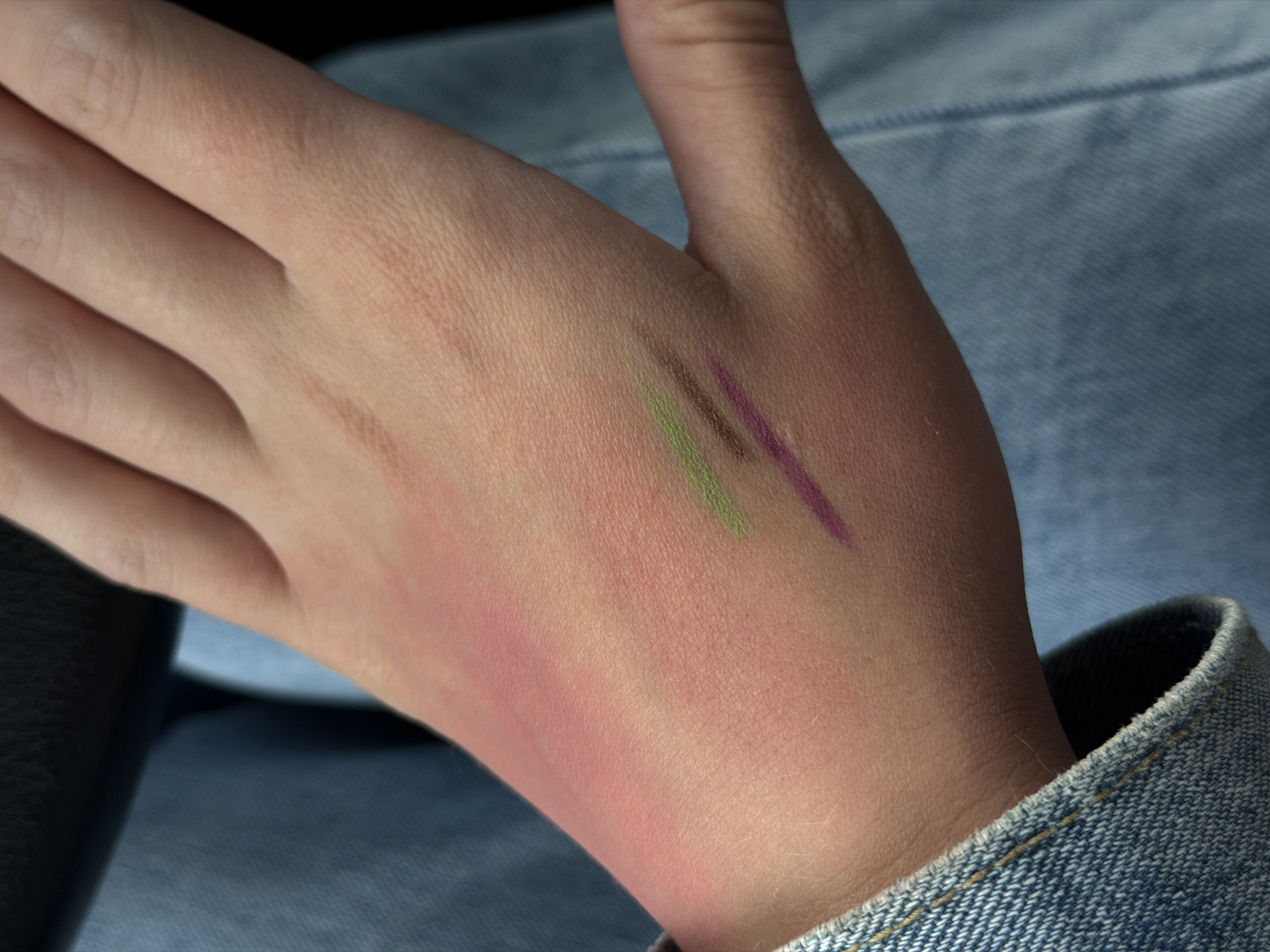 The image presents a close-up view of the back of a person's fair-skinned hand and wrist, likely set in Chaikovsky, Russia, although no specific geographical cues are visible within the frame. The primary focus is on the skin, which displays fine texture and light hair.
The main activity depicted is the swatching of what appear to be cosmetic products. Two distinct, straight lines are drawn across the upper part of the hand: one is an olive green shade, and the other is a rich purple or plum color. These lines are sharp and well-defined, suggesting they were made with makeup pencils or similar applicators, commonly used for testing color and pigment.
Below these swatches, on the lower part of the hand and extending onto the wrist, there is a broader, diffused area of reddish-pink coloration, resembling a blush or a natural flush. This could be another cosmetic product being tested or a natural skin reaction.
In the background and foreground, to the right and bottom right of the hand, a light blue denim fabric is visible, likely part of clothing such as a jacket sleeve or jeans. Its weave and a stitched cuff are discernible, providing a casual context. To the far left, a dark, indistinct object or fabric is partially visible. The lighting is soft and natural, creating subtle shadows that highlight the contours of the hand without indicating a specific time of day or weather conditions. No visible text is present in the image, and no specific emotions are discernible from the close-up of the hand.