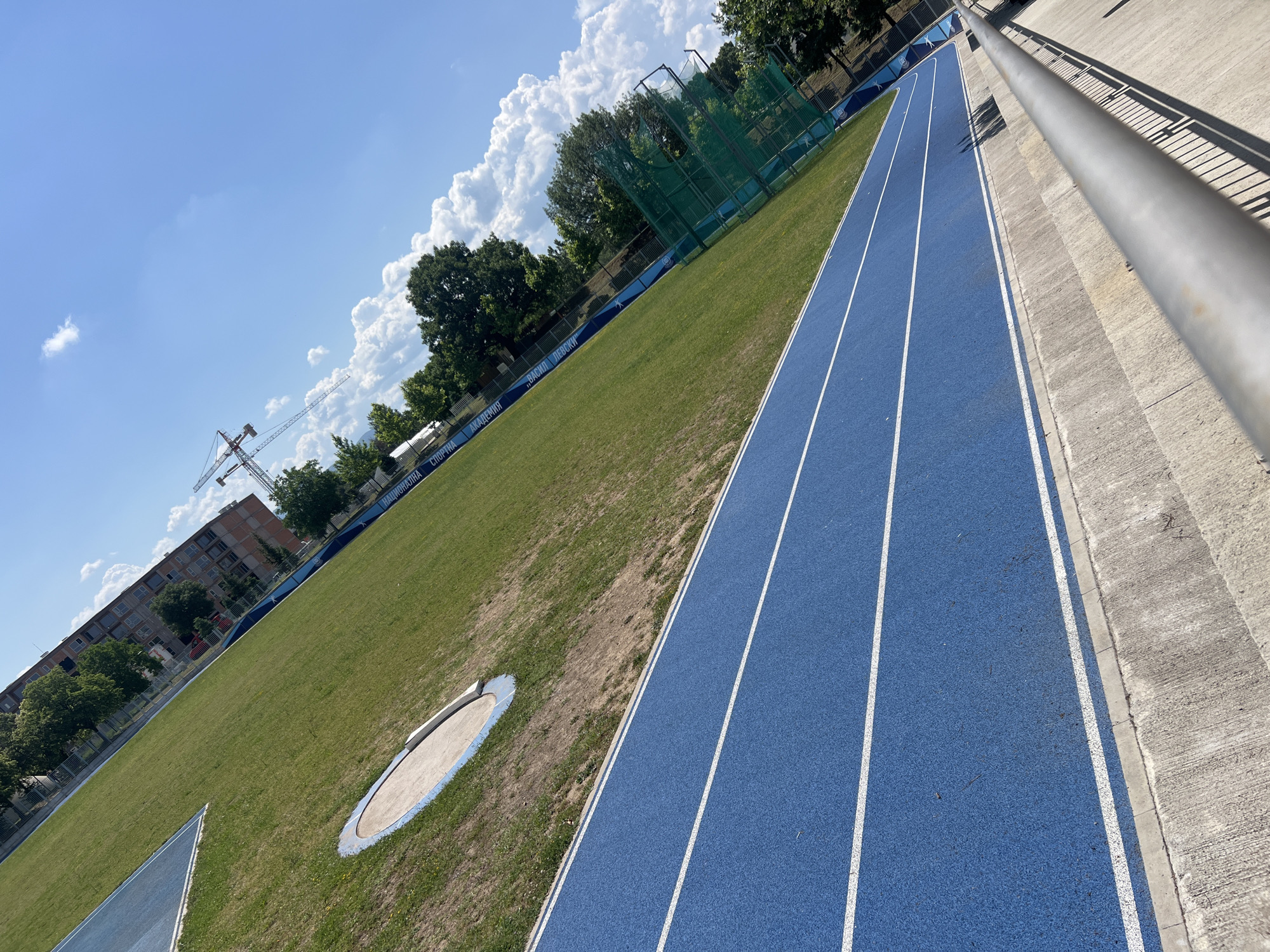 The image depicts an outdoor athletics track with blue lanes and white markings, situated within a grassy field. A circular throwing sector, likely for shot put or discus, is visible on the grass. In the background, a large, multi-story brick building stands, along with trees and a construction crane. A metal railing lines the lower right portion of the frame, suggesting spectator seating. A blue banner with text is visible along the edge of the field. The sky is clear with scattered white clouds. The location is identified as the Vasil Levski National Sports Academy stadium in Sofia, Bulgaria. There are no people or animals present in the image.