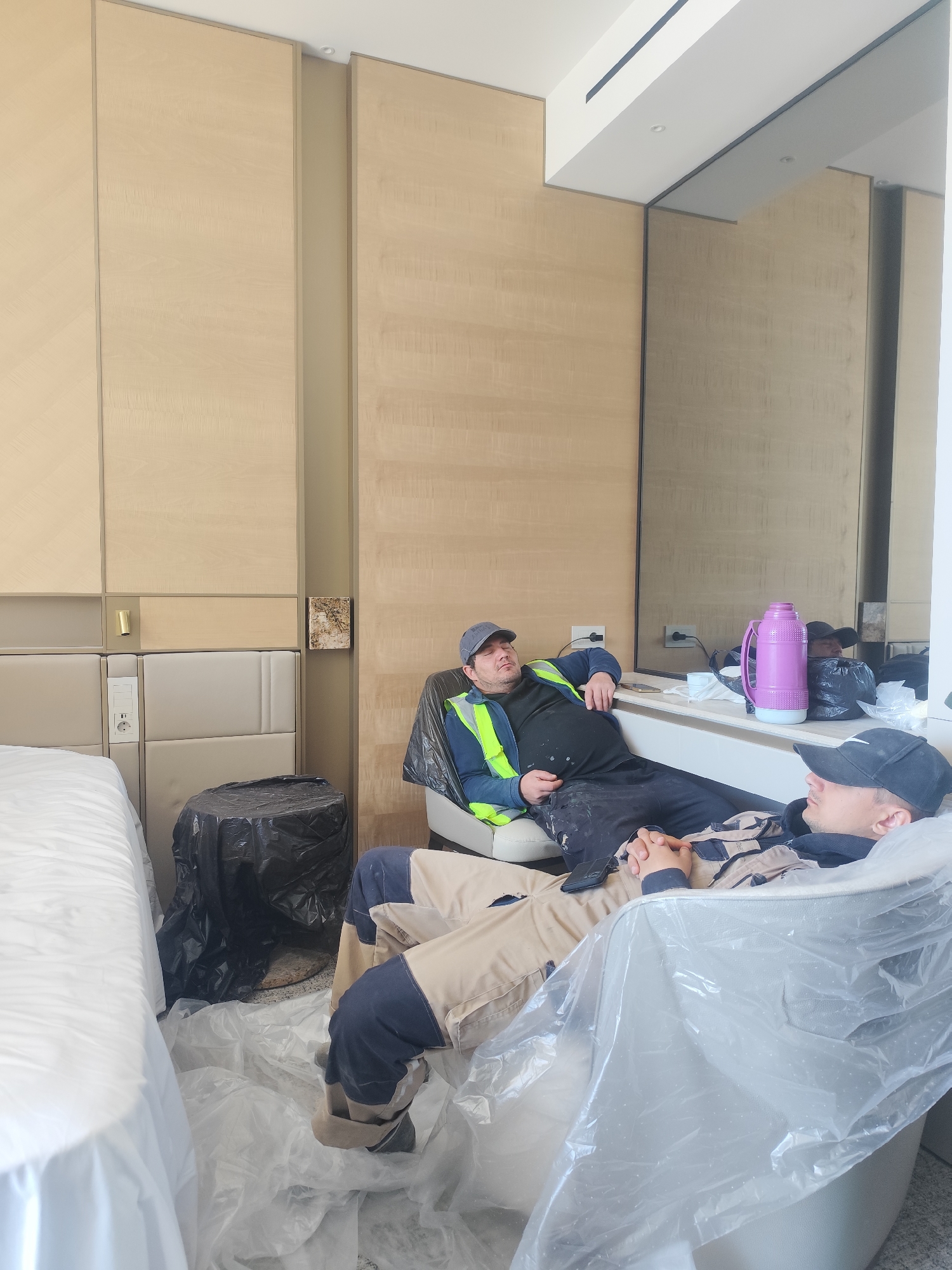 Two construction workers are resting in a hotel room that is undergoing renovations. The room has light brown wood paneling behind the headboard of a bed. The bed is covered in a white sheet. There is a black trash bag on the floor near the bed. To the right of the bed, a worker in a blue hat and a yellow and green safety vest is reclining on a white armchair. He is leaning back against a countertop, where a pink thermos and other items are placed. Another worker, wearing a black baseball cap, is lying on a chair, which is covered in clear plastic. The floor of the room is covered in clear plastic sheeting.