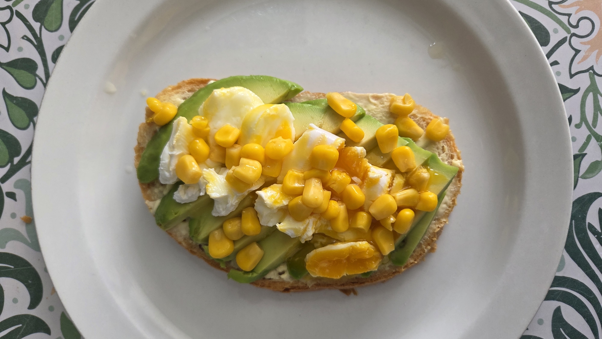 A top-down shot displays a single slice of toast topped with avocado slices, scrambled egg, and corn kernels, served on a white plate. The toast, a light brown hue, forms the base of the dish. Sliced avocado, a vibrant green, is arranged on top of the toast, interspersed with fluffy white and yellow scrambled egg pieces. Yellow corn kernels are scattered generously over the avocado and egg. In the upper left portion of the toast, there are a few thin slices of orange, possibly mandarin orange segments, adding a touch of color. The plate rests on a surface decorated with a green and white leafy pattern. No individuals are visible in the image, and no specific location in Tynset, Norway, can be identified from the provided scene.