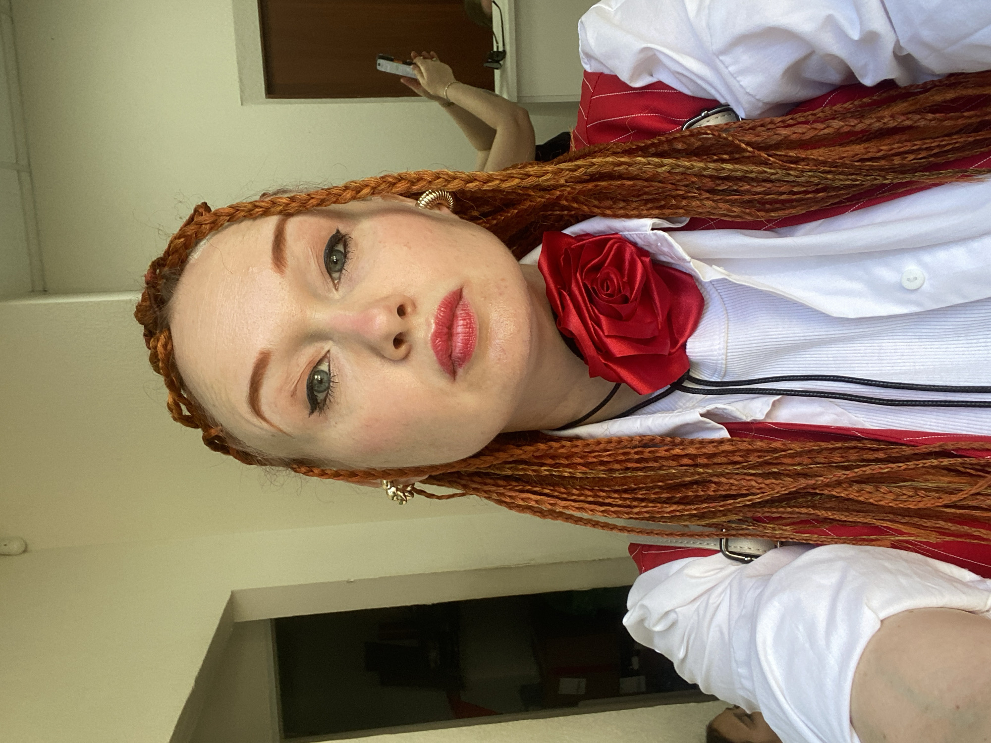 A woman with long, braided, orange hair and bright red lipstick poses for a selfie indoors. She is wearing a white collared shirt, a red vest with pinstripes, and a large red fabric rose pinned to her chest. She has large gold hoop earrings. In the background, a doorway leads to a dark room, and another person is visible in the distance holding a phone, possibly taking a photo. The lighting suggests it is daytime. There are no discernible landmarks or signs that place this image in Sochi, Russia.