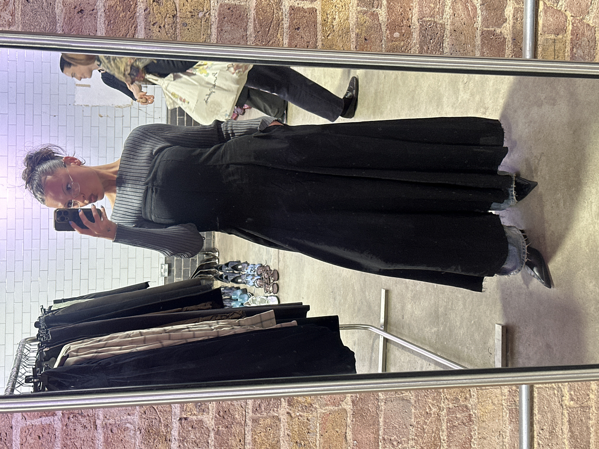 The image depicts a person reflected in a mirror, holding a smartphone, presumably taking a selfie. The individual has their hair up in a bun, wears glasses, a gray striped long-sleeved top, and a long, dark, possibly black skirt. The hem of the skirt appears to have some denim or contrasting material. The person is wearing pointed black shoes.
In the background, reflected in the mirror, there is a clothes rack with various garments hanging on it. Behind the rack and the person, there is a brick wall on one side and a white tiled wall on the other, suggesting an interior space, perhaps a clothing store or fitting room.
Further in the reflection, there's another individual walking by with a patterned bag or tote. Their clothing is darker, and they're wearing black shoes. The floor is a light gray, possibly concrete.
The overall setting appears to be in a retail environment, and the architectural elements of brick and white tile walls are common in many parts of London, United Kingdom, suggesting a possible location in the city.