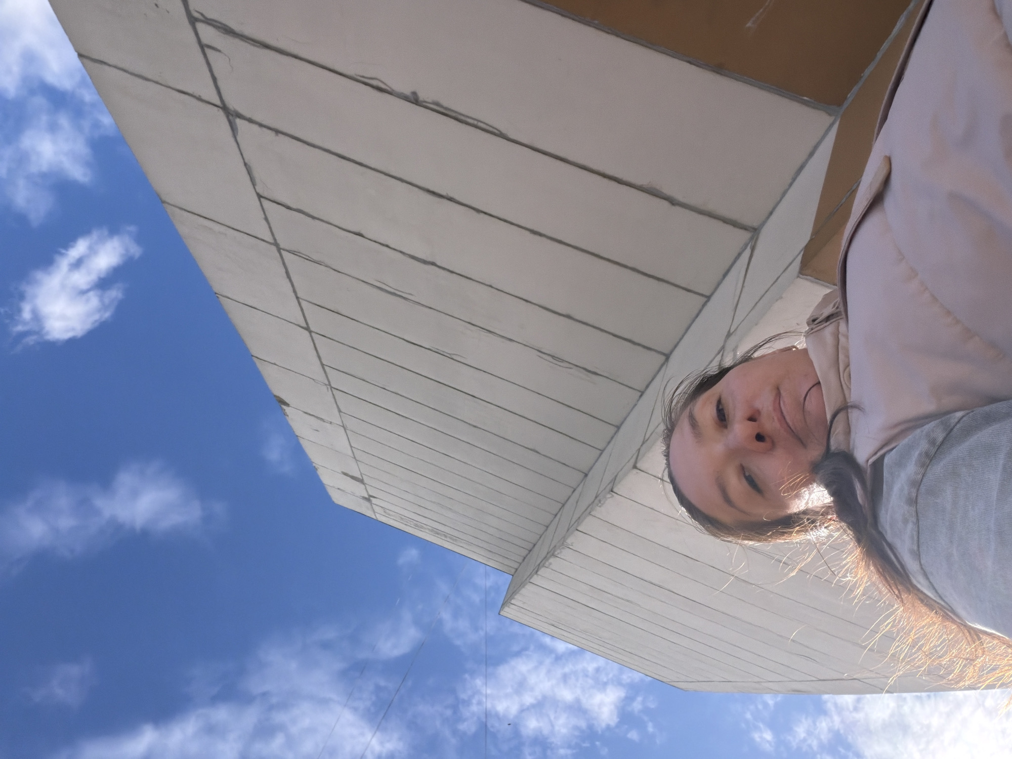 The image is a low-angle shot of a woman looking down at the camera with a building behind her and the sky in the background. The building is white with a linear pattern on its surface. The sky is blue with scattered white clouds. The woman is wearing a light-colored jacket. The city of Staraya is not identifiable in the image.