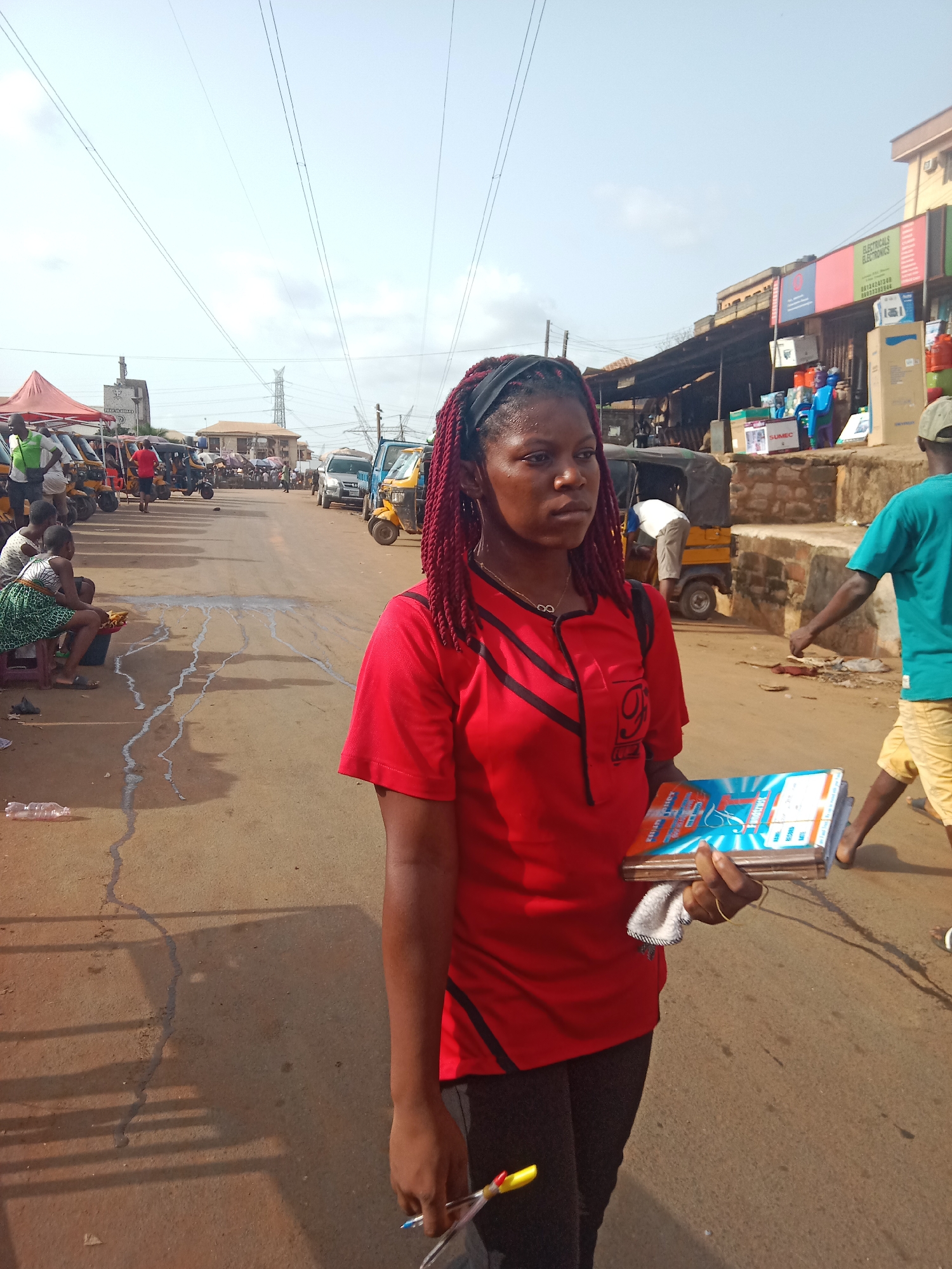In an outdoor scene in Onitsha, Nigeria, a woman with red braided hair stands on a dusty street holding a stack of what appear to be advertising materials. She is wearing a red t-shirt with black trim and black pants. The street shows evidence of heavy use and some staining. In the background, there is a mix of people, parked auto-rickshaws (tuk-tuks), and buildings. A group of people sits to the left, and a man in a blue shirt and yellow shorts walks to the right. Overhead, power lines stretch across the sky. A shop with signage is visible behind a stone wall on the right.