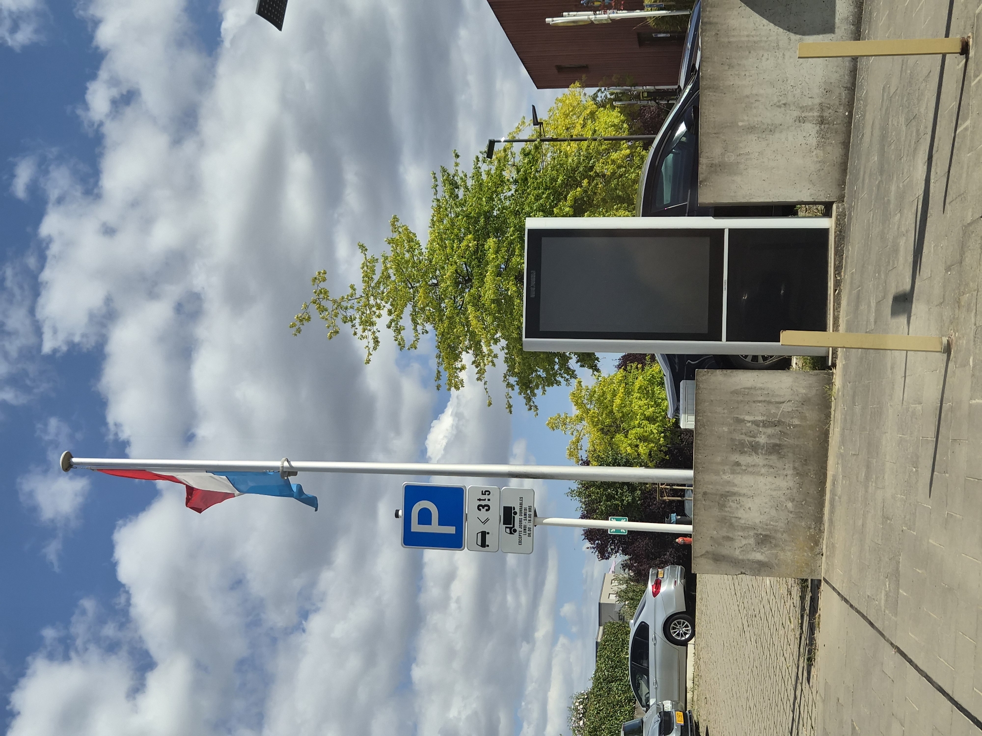 The image shows a public parking area in Mondercange, Luxembourg. A flag of Luxembourg is flying from a flagpole. There are parking signs indicating a parking zone with restrictions. A digital information display is situated near a concrete barrier. Several cars are parked in the area. The environment is outdoors with a cloudy blue sky. The ground is paved with interlocking bricks and concrete slabs. Trees and shrubs are visible in the background. The overall scene depicts a typical urban outdoor space.