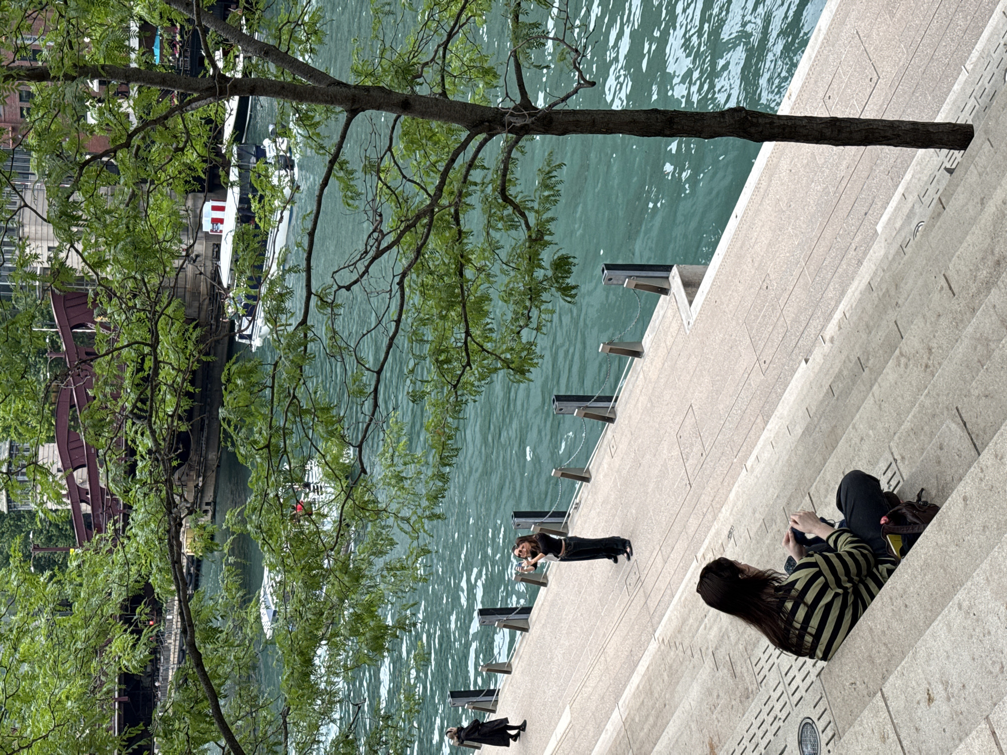 The image depicts a view of the Chicago River in Chicago, Illinois, United States of America. In the foreground, a person sits on concrete steps facing away from the camera, wearing a striped long-sleeved shirt and dark pants, and appears to be looking at a phone. Behind them, a paved walkway runs alongside the river. Two people are standing on the walkway, one in a black outfit taking a picture with a phone, and another to their right. Further in the background, a boat is moving on the turquoise water. A bridge with a red railing is visible in the distance, partially obscured by trees with green foliage. The scene captures a portion of the urban landscape along the riverfront.