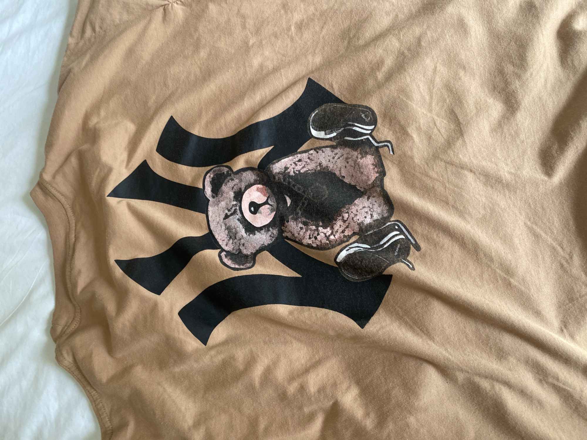 The image shows a close-up, high-angle view of a tan t-shirt laid flat on a white surface, likely a bed or a clean cloth. The t-shirt features a graphic print of a teddy bear sitting in front of a black logo resembling the New York Yankees logo. The teddy bear is rendered in shades of brown and pink, with detailed fur texture. It is wearing what appears to be black and white striped socks or shoes. The t-shirt itself is slightly creased and wrinkled, indicating it is made of a soft fabric like cotton. The background is dominated by the white surface, which is also wrinkled. There is no discernible location in Kuta Selatan, Indonesia, or any other city in the image.