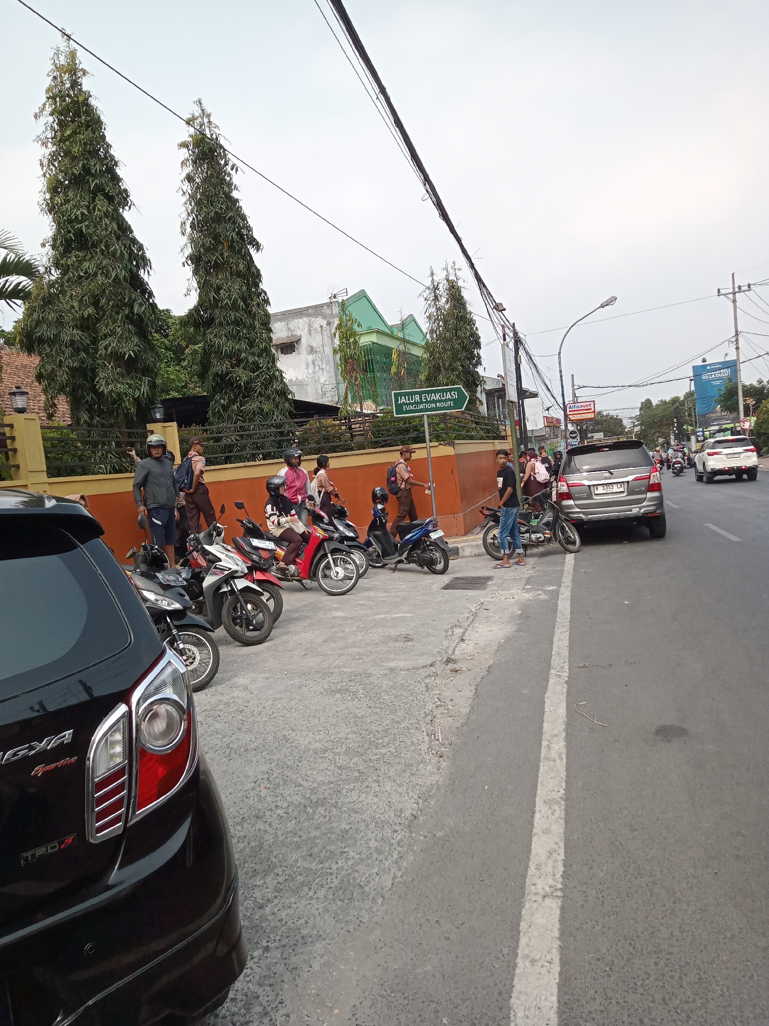 In Batu, Indonesia, a street scene unfolds with several motorbikes parked on a paved area beside a road. A black car is parked in the foreground, partially obscuring the view. To the right, a road with white lane markings is visible, with traffic including a silver SUV and other vehicles in the distance. On the left, a group of people are gathered around the parked motorbikes, some standing and some seated on them. One man in a gray jacket and blue shorts stands near a motorbike, holding a helmet. Behind them, a building with a brown fence and trees can be seen. A green sign indicating "JALUR EVAKUASI" (Evacuation Route) is mounted on a pole. The sky is overcast.