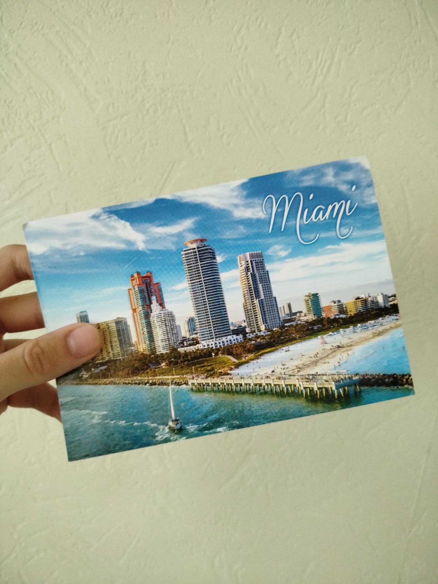 A hand is holding a postcard featuring an image of the city of Miami, Florida. The postcard displays a view of Miami's skyline, including several skyscrapers, a pier extending into the water, a beach with people, and a sailboat. The word "Miami" is written in cursive at the top right of the card. The background behind the hand and postcard appears to be a plain, textured wall.