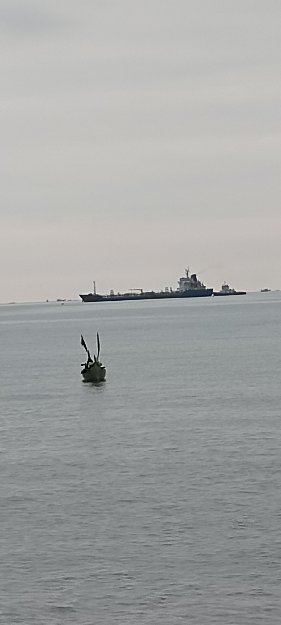 In a wide shot of the sea, a small, weathered boat with two upright poles that may have once held sails or flags floats in the foreground. The boat appears empty. In the middle ground, a long cargo ship, likely a tanker, is visible. It is being assisted by a smaller tugboat positioned near its stern. The background is a hazy sky with a faint horizon line. The overall scene suggests a calm, overcast day at sea, likely near a port or shipping lane. While the image does not explicitly depict Lhokseumawe, Indonesia, the presence of cargo ships and a port environment are common in coastal cities in Indonesia.