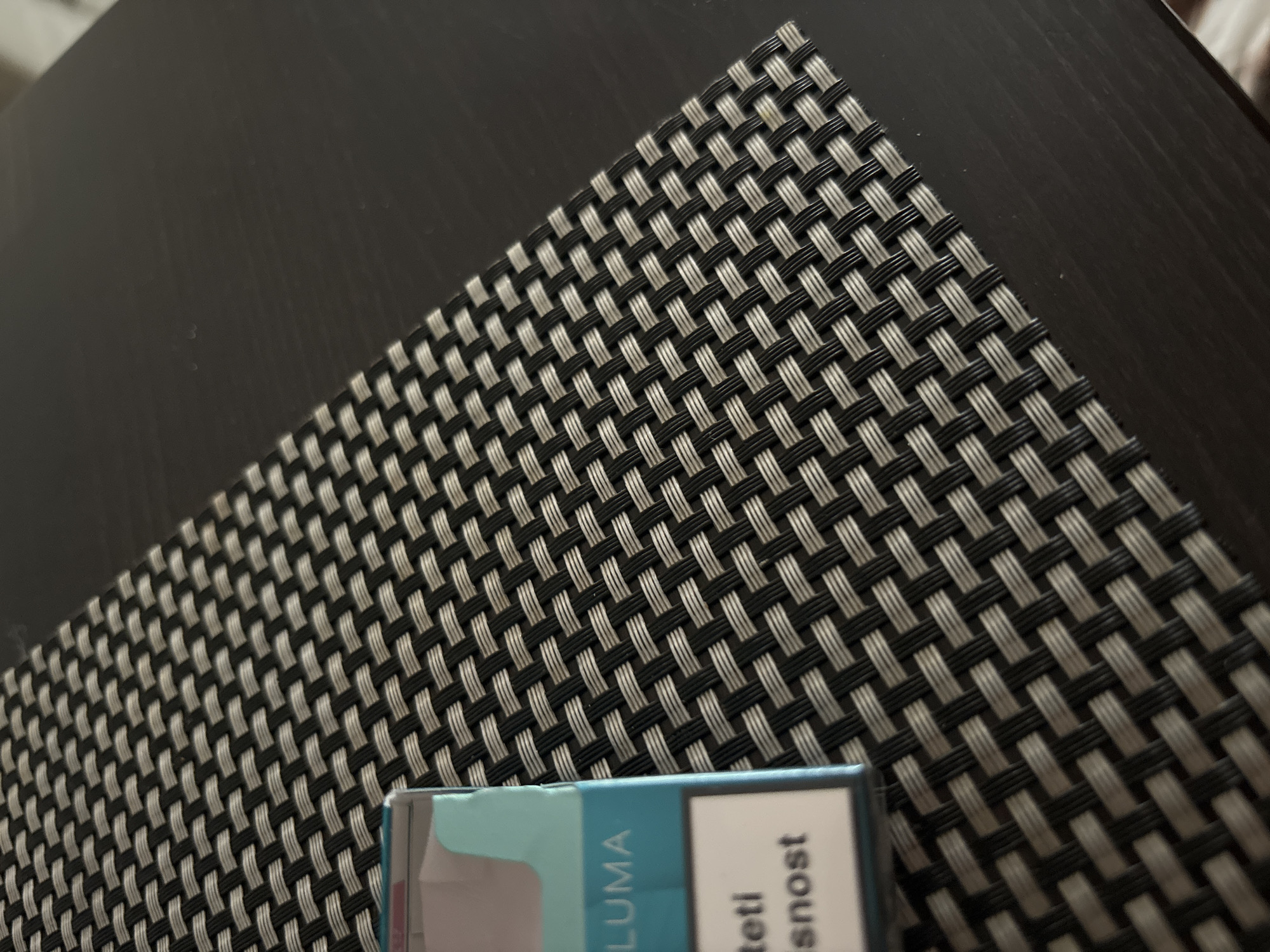 The image shows a close-up view of a woven placemat on a dark-colored table. A partial pack of cigarettes is visible in the lower portion of the frame. The brand name "Luma" is visible on the pack. The words "teti" and "isnost" are also legible on the cigarette pack. The exact location within Budva, Montenegro is not discernable from the provided image.