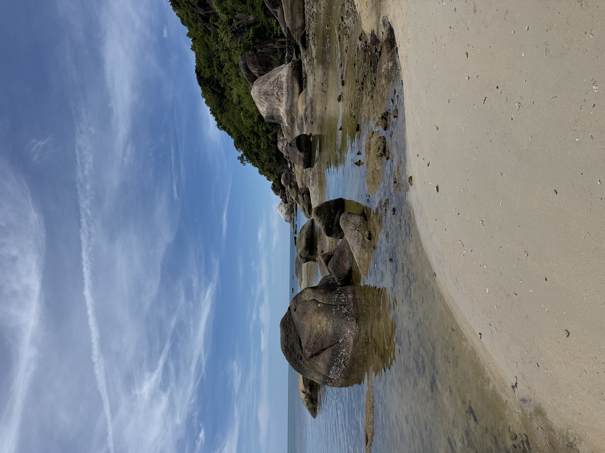 The image depicts a coastal scene, likely a beach, in Baan Chaweng Noi, Thailand. The foreground shows a sandy beach with small debris scattered. Adjacent to the sand is a shallow body of water with several large, dark-colored rocks protruding from it. Some rocks are partially submerged. Beyond the rocks and water is the ocean, appearing calm. In the background, there's a rocky outcrop covered with dense green vegetation. The sky above is light blue with wispy white clouds.