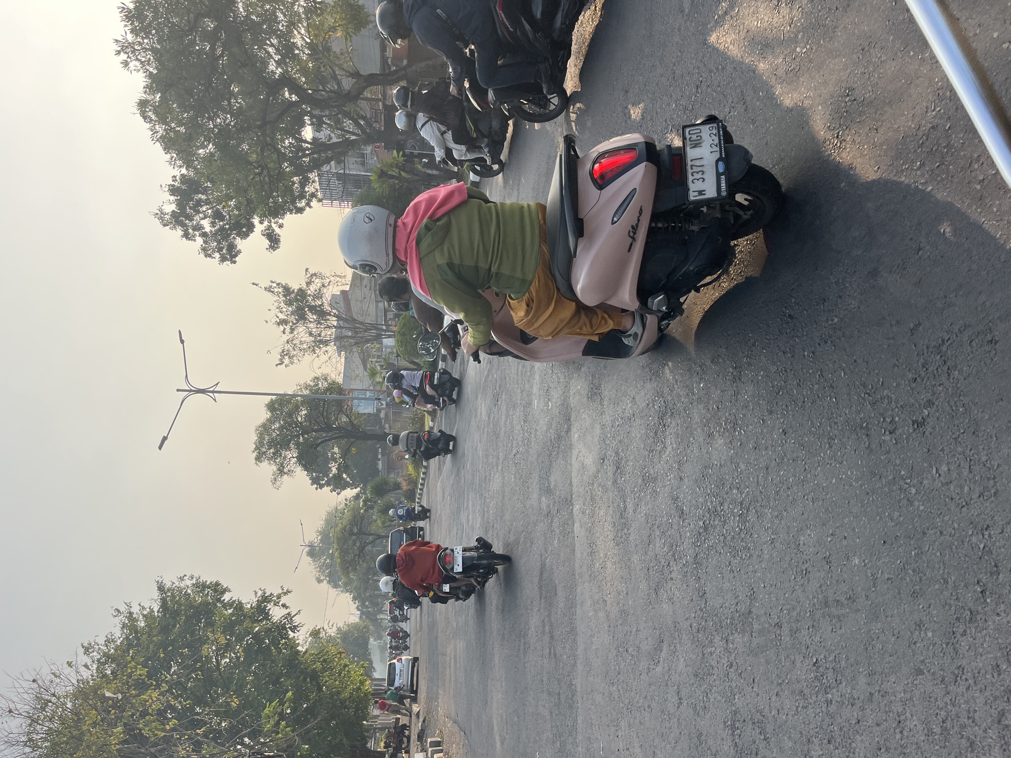 The image depicts a street scene in Tenggulunan, Indonesia, during daylight. A line of motorbikes is traveling down a paved road lined with trees and some buildings in the background. In the foreground, a person wearing a pink scarf and a green jacket rides a pink motorbike. The motorbike has a license plate that reads "W 3371 NGD 12-29". Several other motorbikes with riders, many wearing helmets, are visible in front of and behind this central motorbike, suggesting a moderate flow of traffic. The sky is bright, indicating it is either morning or late afternoon, with the sunlight casting shadows on the road. The overall impression is of a typical urban or suburban street with a prevalence of motorcycle transportation.
