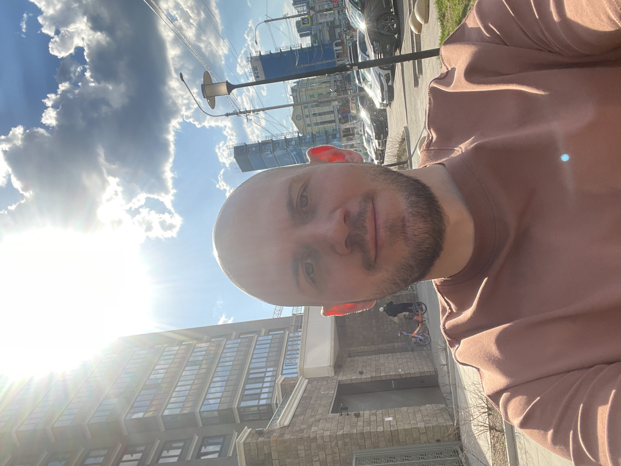 The image shows a man with a shaved head and a short beard taking a selfie. He is wearing a brown shirt. The background shows a street in Krasnoyarsk, Russia. A building with many windows is on the left side of the image. Streetlights and power lines are also visible. Several cars are parked along the street. A construction crane is visible in the distance. A bicycle is also visible in the lower center of the image. The sky is blue with white clouds, and the sun is shining brightly.