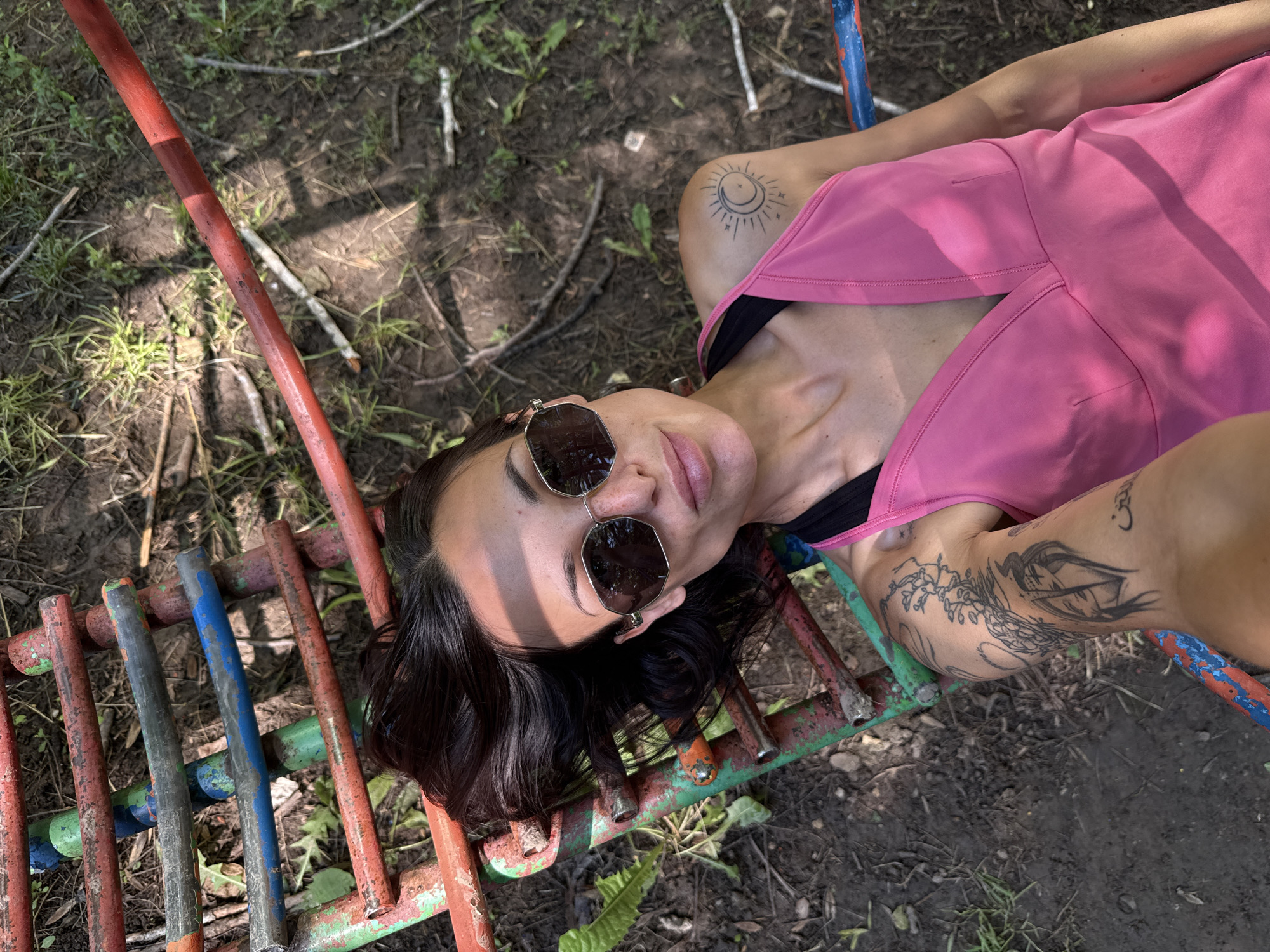 A young woman is captured lying on her back, looking upwards towards the camera. She has dark hair and wears large, reflective sunglasses that conceal her eyes, with her lips slightly parted. She is dressed in a bright pink sleeveless top, layered over a black top or bra.
Her head and body are supported by an old, multi-colored metal structure, appearing to be part of playground equipment, with bars painted in faded reds, blues, and greens. The ground beneath and surrounding her is a mix of dirt, sparse green grass, and scattered dry sticks and leaves, indicating an outdoor, somewhat natural or unkempt setting.
Notable tattoos are visible on her arms. On her right shoulder, there is a design featuring a sun and a crescent moon. Her left arm displays a larger, intricate tattoo that appears to be a winged figure or mythical creature intertwined with floral patterns. A small Om symbol is also visible on her upper right arm.
The scene is illuminated by strong natural sunlight, casting distinct shadows across her body and the ground, suggesting a clear, bright day. The photograph is taken from an overhead perspective. The overall context suggests a relaxed outdoor moment, potentially in a park or recreational area in Pleven, Bulgaria. No visible text is present in the image.