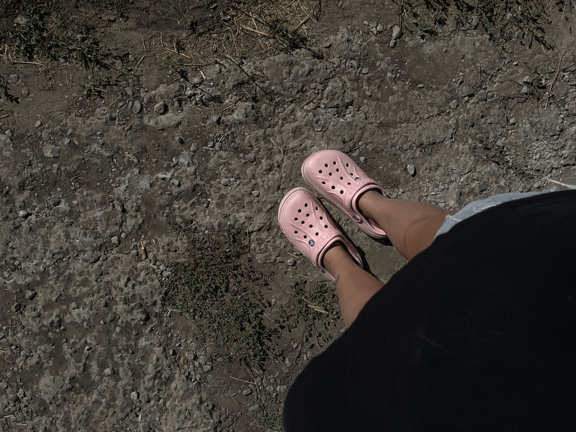 A person is standing on a dirt and gravel path. The person is wearing light pink Croc shoes, denim shorts, and a dark top that covers their torso and left leg. The shoes are the primary focus of the image, with their distinctive perforated design clearly visible. The ground is rough and uneven, with small stones and patches of dry grass. The scene appears to be outdoors on a sunny day, casting a distinct shadow from the person onto the ground. There is no specific information to suggest the location is Rostov-on-Don, Russia.