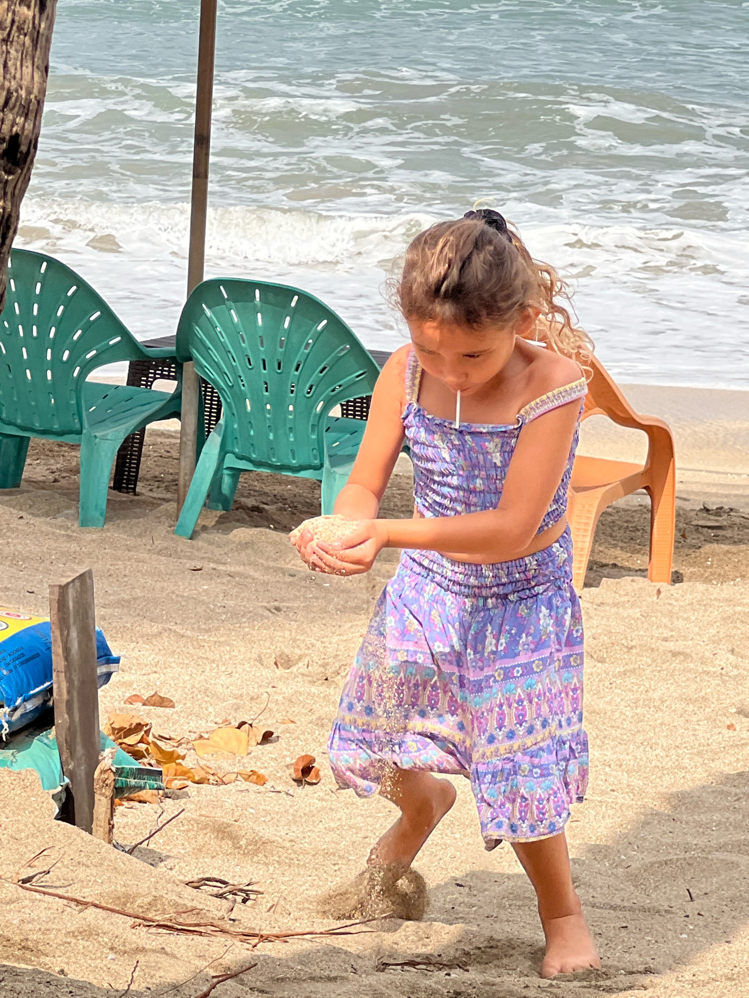 A young girl with blonde, curly hair is playing on a sandy beach. She is wearing a purple floral print two-piece outfit. Her hands are cupped, holding a small amount of sand. She is looking down at the sand in her hands. In the background, the ocean waves are breaking on the shore, and several green plastic beach chairs and one orange plastic chair are visible. A wooden pole, possibly part of a beach umbrella, stands near the girl. The overall scene is set on a sunny day on the beach in Kuta, Indonesia.