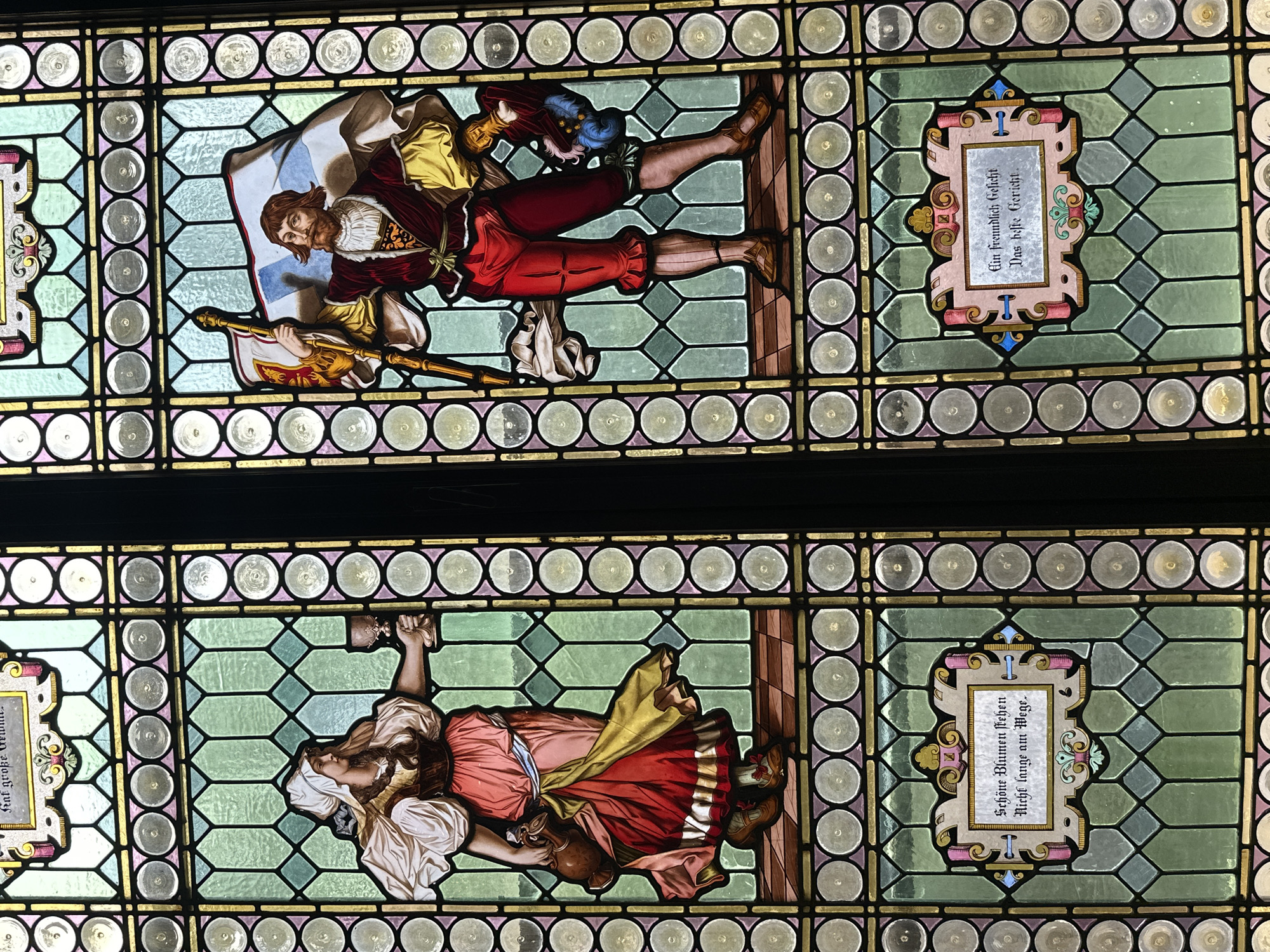 The image shows two stained glass windows. The upper window depicts a man in medieval clothing, holding a flag and a staff. He wears red pants, a maroon jacket, and a white shirt. The flag has a blue and white design. The bottom window features a woman in similar attire, holding a hammer. She wears a red and white dress with a yellow sash. The background of both windows consists of geometric patterns in green and gold. Adjacent to each figure is a framed inscription in German. These stained glass windows are not located in Tlalnepantla, Mexico.
