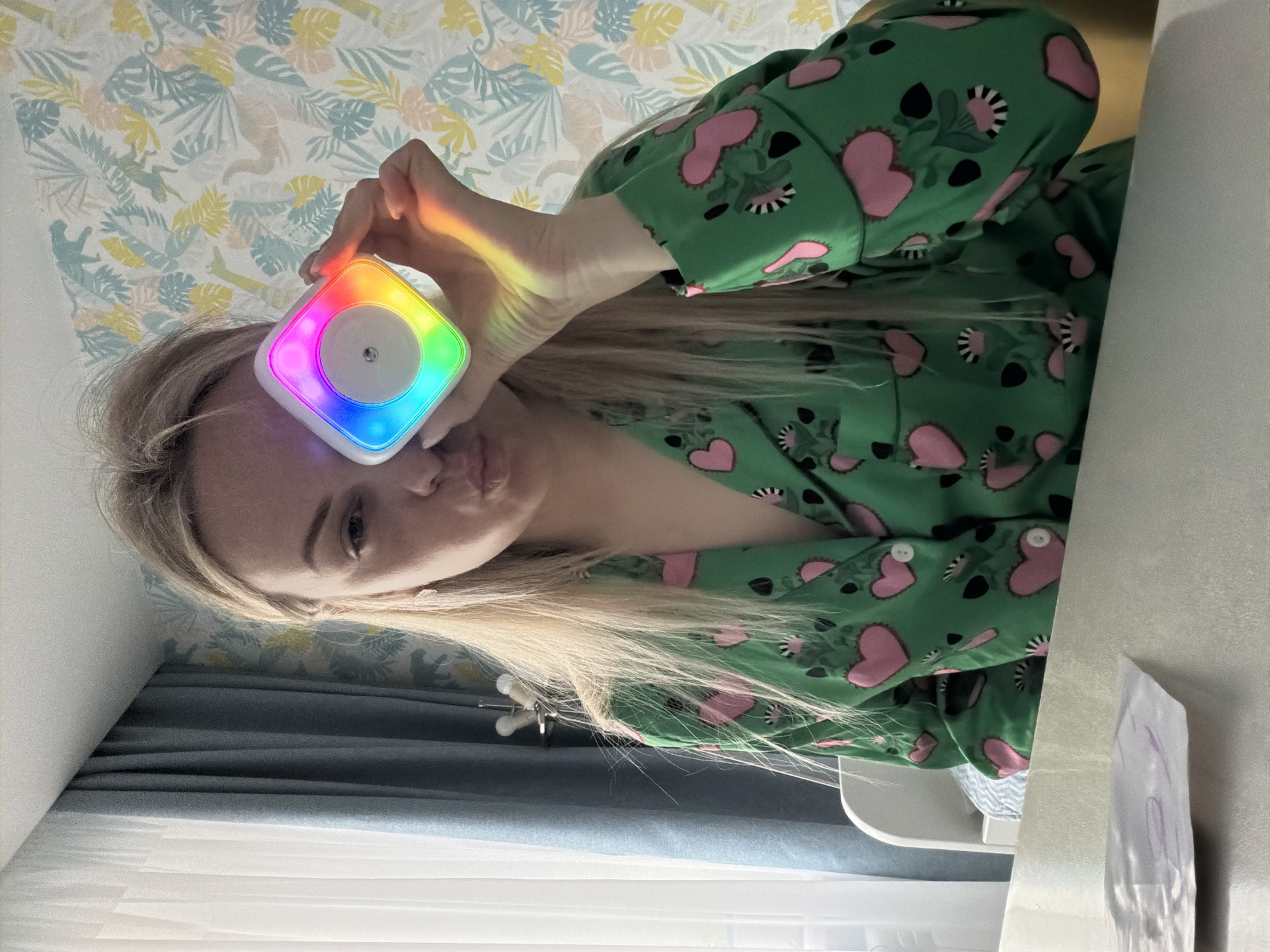 A portrait captures a young woman in an indoor setting. She has long, blonde hair and is wearing green long-sleeved pajamas patterned with pink hearts, black shapes, and white flowers. She holds a small, square, white electronic device with a glowing rainbow-colored edge close to her face, casting a colorful light onto her fingers. Her face is partially visible, displaying one eye and a slightly pursed-lip expression as she looks towards the viewer.
The background features a wall covered in light-toned wallpaper adorned with a repeating pattern of light blue, yellow, and green tropical leaves and abstract animal figures. Below her arm, dark blue curtains are visible. To the right of the frame, a light-colored surface, possibly a table, holds a white bag with faint, illegible purple writing. A small metal tripod or stand is also partially visible near her elbow. The scene conveys a relaxed, home atmosphere, likely a bedroom, with the illuminated device suggesting evening or nighttime activity, or simply a playful moment. The location context for this indoor scene is Votkinsk, Russia.