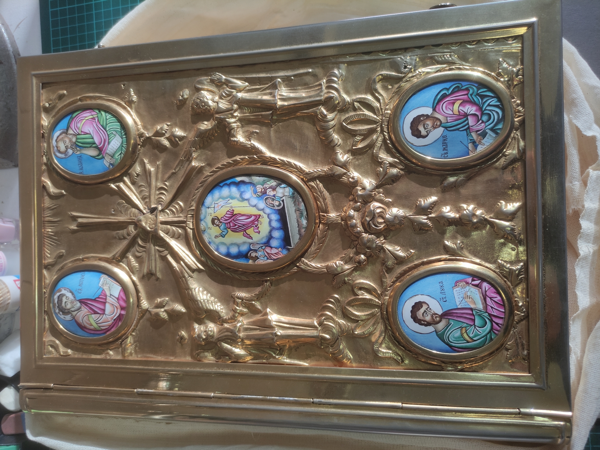 An ornate, gold-colored, metallic book cover, possibly brass or gold-plated, is depicted from a slightly elevated perspective. The cover features intricate relief sculpture and five inset, painted enamel or icon-style medallions. The central oval medallion illustrates the Ascension of Christ, showing a robed figure ascending towards the sky with smaller figures below looking upwards. Surrounding the central medallion are four winged angelic figures in relief, positioned vertically, and detailed floral and scrollwork patterns. Four additional oval medallions are situated at each corner of the cover. Clockwise from the top left, these depict: a bearded male figure, identified in Cyrillic as "Св. Матθеи" (St. Matthew), writing in a book; a bearded male figure, identified as "Р. Iωаннъ" (St. John), in contemplation with a scroll; a bearded male figure, identified as "Св. Марко" (St. Mark), writing; and a bearded male figure, identified as "Ст. Лука" (St. Luke), also writing. The entire cover has a raised border, and a section resembling a book spine is visible on the left edge. The object rests on a light-colored, textured fabric surface, with blurred objects and a green grid cutting mat visible in the background, suggesting an indoor setting. The location in the city of Moscow, Russia, cannot be determined from the provided visual information.