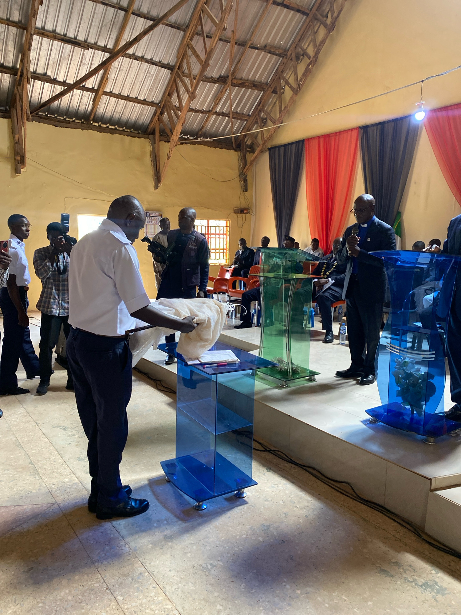 The image depicts an indoor gathering, likely a church service or similar event, in Ohit, Nigeria. In the foreground, a man in a white shirt and dark trousers is presenting a white cloth, possibly a religious artifact, to another man dressed in a suit with a clerical collar. Behind them, several individuals are observing, with one person holding a camera and another a video recorder. The environment features a high, exposed wooden and corrugated metal roof. The background is decorated with red and grey curtains. Several modern, clear acrylic podiums are visible, two of which are a striking blue color. The floor is tiled with a speckled pattern. In the middle ground, a man in a suit and clerical collar stands at a podium, holding a microphone, suggesting he is speaking. He is surrounded by other men in similar attire, seated or standing, some of whom appear to be clergy. The lighting suggests an indoor setting, possibly illuminated by a single bare bulb hanging from a wire and natural light from windows, though the windows are not clearly visible. The overall atmosphere appears formal and solemn.