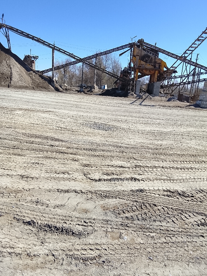 This image depicts an active or disused industrial aggregate processing facility under a clear blue sky in Yangi Chinoz, Uzbekistan. The foreground consists of an expansive, unpaved dirt and gravel ground heavily marked with numerous tire tracks, indicating vehicular traffic. In the midground and background, a complex system of industrial machinery is visible. This includes multiple long, dark-colored conveyor belts supported by lattice metal structures, extending horizontally across the scene. To the left, a large pile of dark brown earth or aggregate rises. To the right, a prominent, large yellow industrial processing unit, possibly a screener or crusher, is centrally located within the conveyor system. Scattered smaller piles of material are visible beneath and around this machinery. Bare deciduous trees are present in the distant background, beyond the industrial equipment. No individuals are visible within the scene, and no overt actions or interactions are depicted. The overall environment suggests an industrial site for material handling.