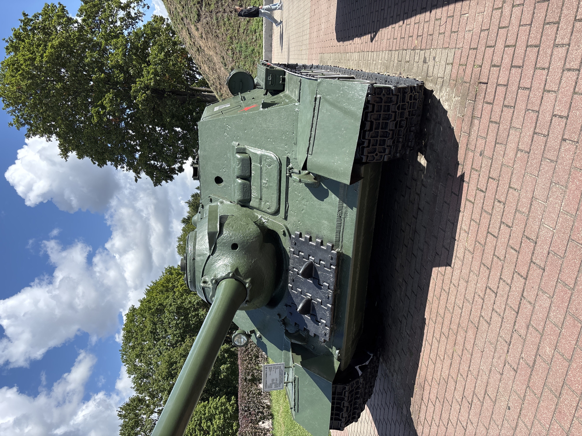 The image shows a Soviet SU-100 tank destroyer on display at the Brest Fortress in Brest, Belarus. The tank is painted green and is positioned on a red brick paved area. In the background, there are trees and a blue sky with white clouds. A person is walking away from the tank in the middle ground on the right side of the image. A small informational plaque is visible on the front left of the tank.