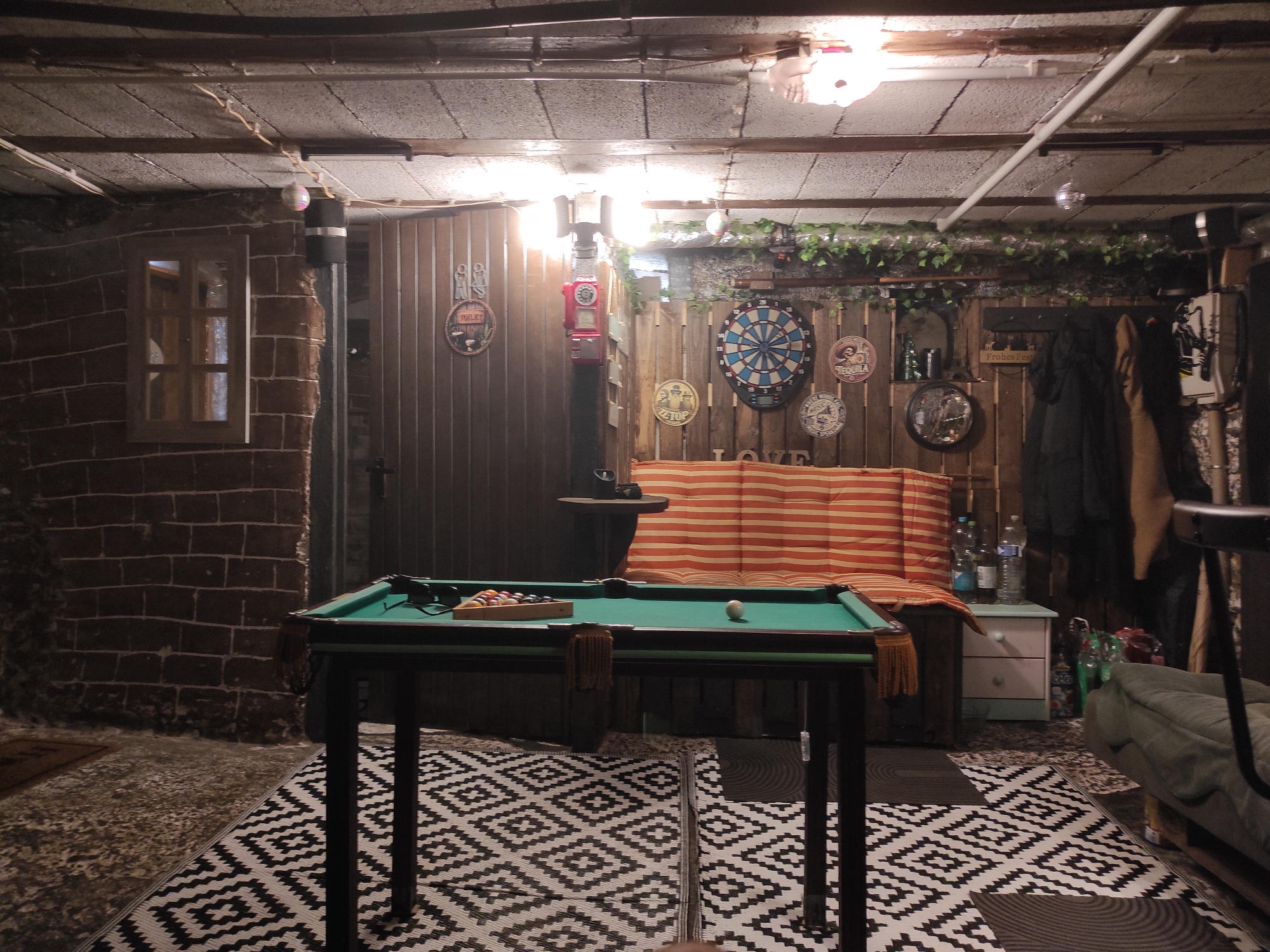 This is a dimly lit, indoor recreation room, possibly a basement or garage, in Waldbronn, Germany. The room features a pool table with billiard balls racked and one ball on the felt. A patterned sofa with orange and white stripes is visible behind the pool table, along with a small white chest of drawers. The walls are constructed of brick and wood paneling, decorated with various circular signs and a dartboard. A red vintage gas pump is mounted on the wall. A window with wooden framing is set into the brick wall. The ceiling is made of concrete tiles and exposed beams, with overhead lighting and some decorative string lights. There is a patterned rug on the floor. A green sofa is partially visible in the lower right corner. Clothing items are hanging on the right side of the room. The overall atmosphere is that of a casual, rustic hangout space.