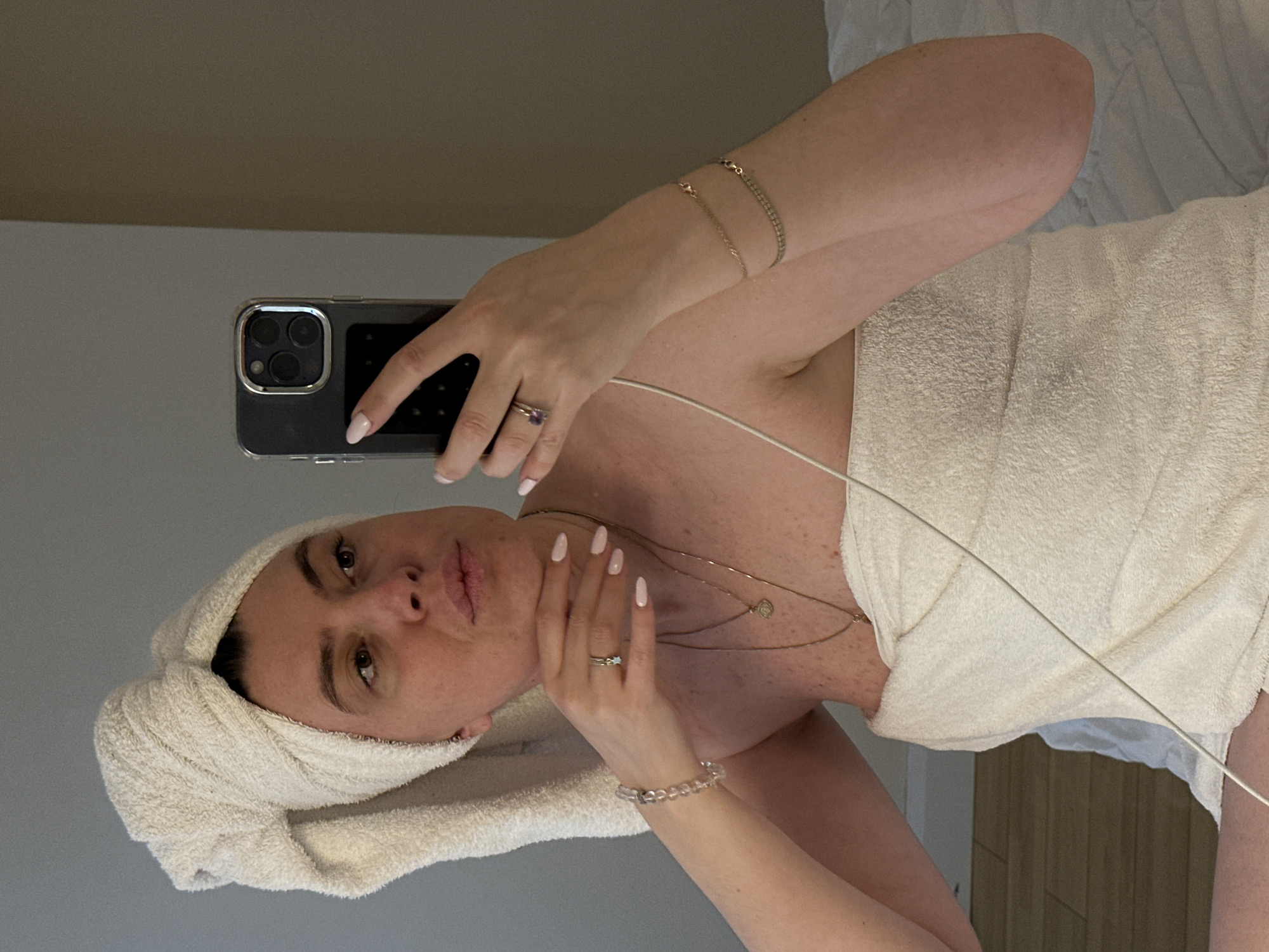 A woman is taking a selfie in a mirror. She is wearing a white towel wrapped around her hair and body. She is holding an iPhone in her right hand. She is wearing several pieces of jewelry including rings, necklaces, and bracelets. The background is a neutral grey color. The location is not discernible from the image.