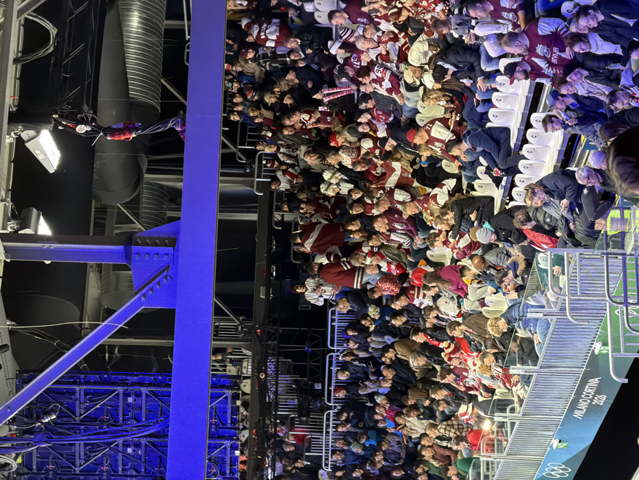 A large crowd fills stadium seating, many wearing maroon and white team colors. To the left, structural elements are visible, including blue painted support beams, pipes, and lighting fixtures. Signage for "MILANO CORTINA 2026" is partially visible in the lower right corner. The overall composition suggests a sporting event or large gathering at an indoor arena.