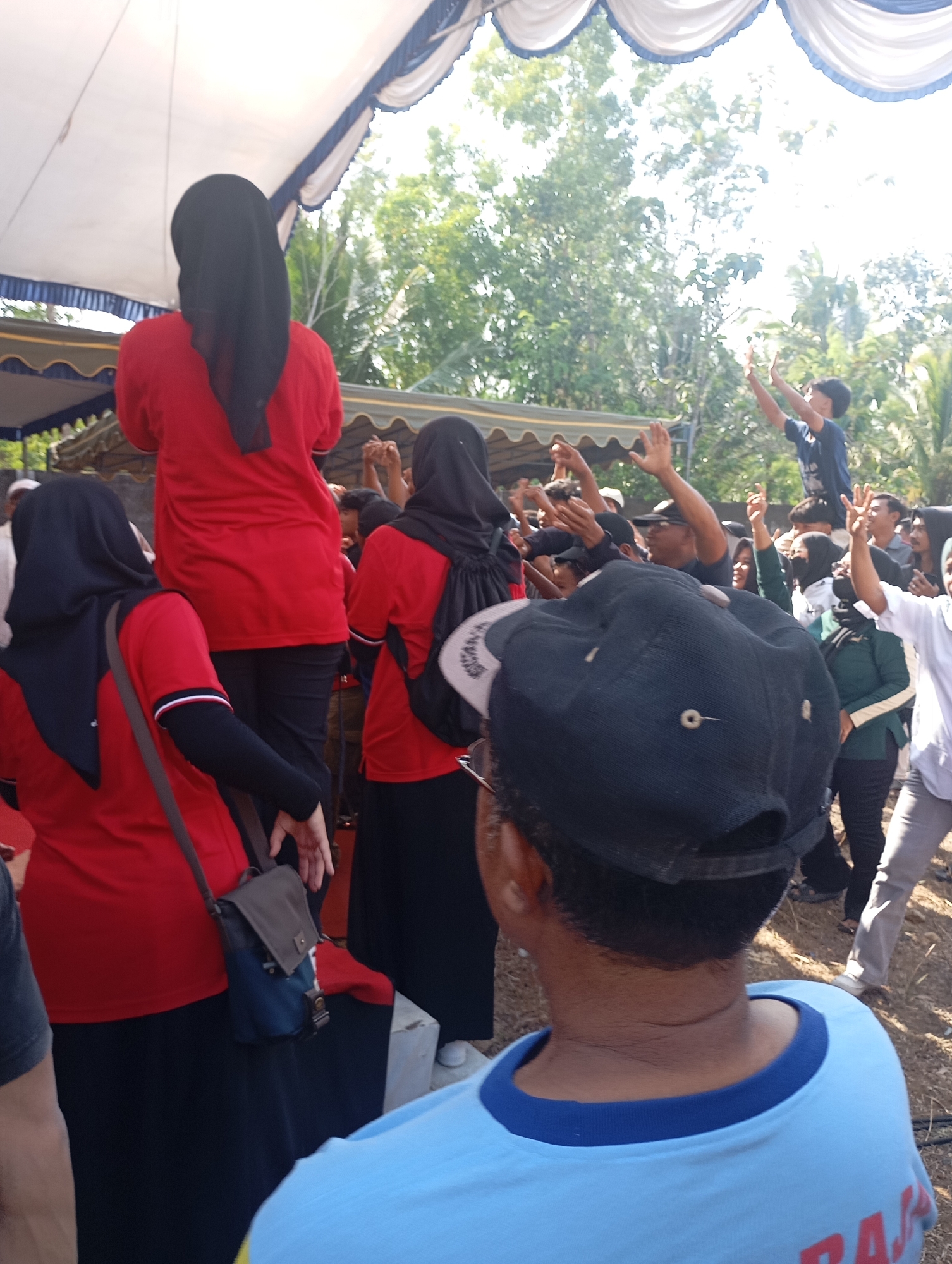 The image depicts an outdoor gathering under a tent in Ketepung, Indonesia. Several people are visible, many wearing red shirts and black hijabs. In the foreground, the back of a person wearing a light blue t-shirt and a black baseball cap is prominent. They appear to be looking towards a crowd. To the left, a woman in a red shirt and black hijab is looking downwards, and a messenger bag is slung across her body. Behind her, another woman in a red shirt and black hijab is standing. Further back, a group of people are gathered, with some raising their hands. One young person in a dark blue t-shirt is visible in the upper right portion of the crowd, with their hands raised. The environment appears to be somewhat rural, with trees visible in the background and a dirt or gravel ground. The lighting suggests it is daytime, possibly sunny, with some areas of bright light and shadows.