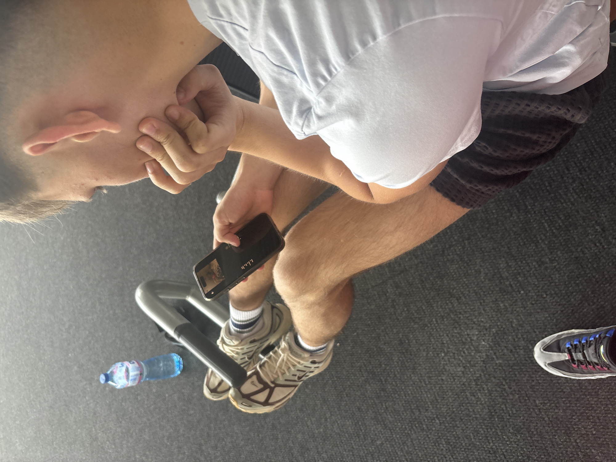 The image captures an indoor scene, likely a gym or fitness facility in Blagoevgrad, Bulgaria. A person, appearing male, is seated, viewed from a high angle. They wear a light-colored t-shirt, dark, textured shorts, and beige athletic shoes with white socks. Their left hand is raised towards their ear or cheek, while their right hand holds a black smartphone. The phone case features the Cyrillic text "УШИ" (Ushi, meaning "ears"). The phone screen displays an image, possibly a video or photo.
To the left of the person's feet, a blue plastic water bottle with a blue cap lies on the dark, carpeted floor. The bottle's label clearly shows the brand "DEVIN" and "Изворна вода" (Spring Water), indicating a Bulgarian product. Part of a silver and black gym equipment bar is visible beneath the seated person. In the bottom right corner, the toe of another athletic shoe, grey with black, pink, and blue laces, suggests the presence of another individual or footwear placed nearby. The overall impression is that of someone resting or taking a break from a workout.