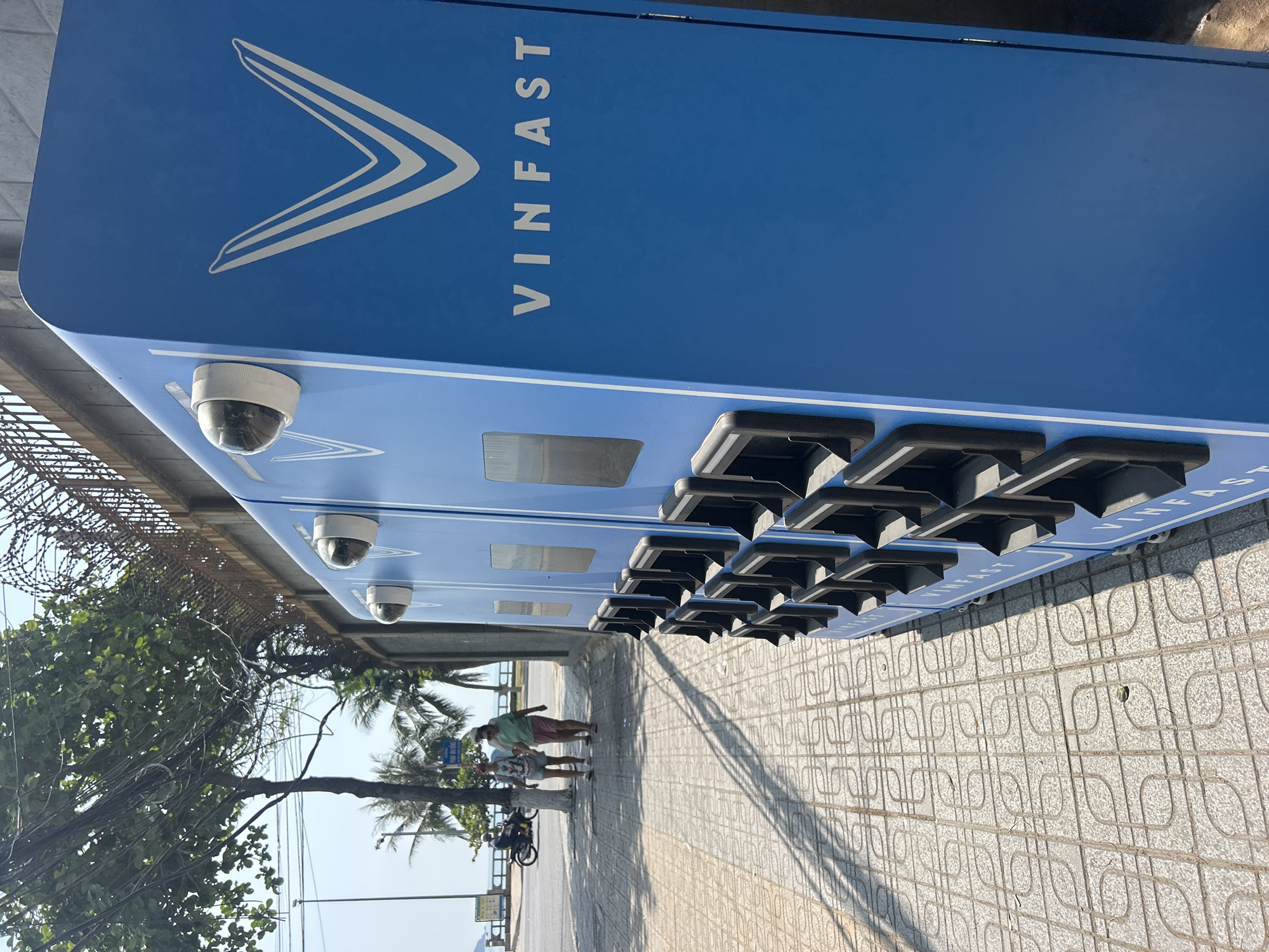 The image depicts an outdoor scene in Bắc Nha Trang, Vietnam, on a bright, sunny day, suggested by the clear sky and pronounced shadows. A large, blue battery swapping station, prominently featuring the "VINFAST" text and its distinctive V-shaped logo in white, occupies the left side of the frame. The station appears to be a tall, rectangular unit with several slots for batteries arranged in rows, though no batteries are currently visible in the slots. Two dome-style surveillance cameras are mounted on its side, one near the top and another lower down.
To the right of the station, a paved sidewalk with a repeating patterned tile design stretches into the distance. Two individuals are walking on the sidewalk, with one possibly on a bicycle further down. Lush green trees, including palm trees, line the sidewalk, providing some shade. In the upper left background, behind the battery station, a metal structure topped with barbed wire is visible. The bright sunlight casts long shadows of the battery station and trees across the sidewalk, indicating it is likely mid-morning or afternoon. The overall impression is one of a modern urban street environment.