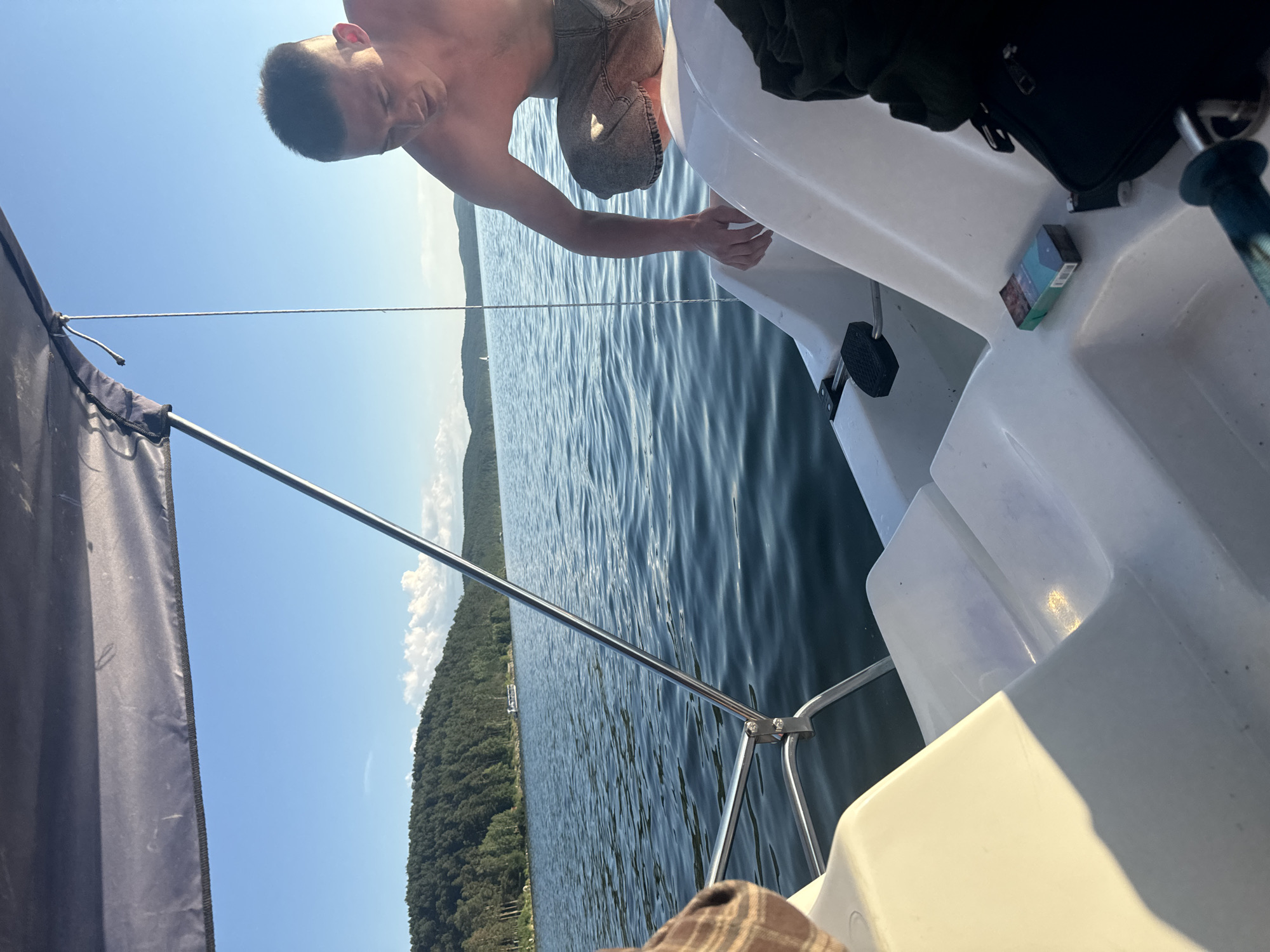 A young shirtless man in denim shorts is crouching on a white boat on the Turgoyak Lake in Turgoyak, Russia. He is reaching into the boat as if to retrieve something. The lake is calm with small ripples and surrounded by lush green forested hills. The sky is a clear blue with scattered white clouds. The boat has a dark fabric canopy providing shade. A pack of cigarettes and a lighter are visible on the boat's surface. The overall scene depicts a leisurely activity on a body of water on a sunny day.