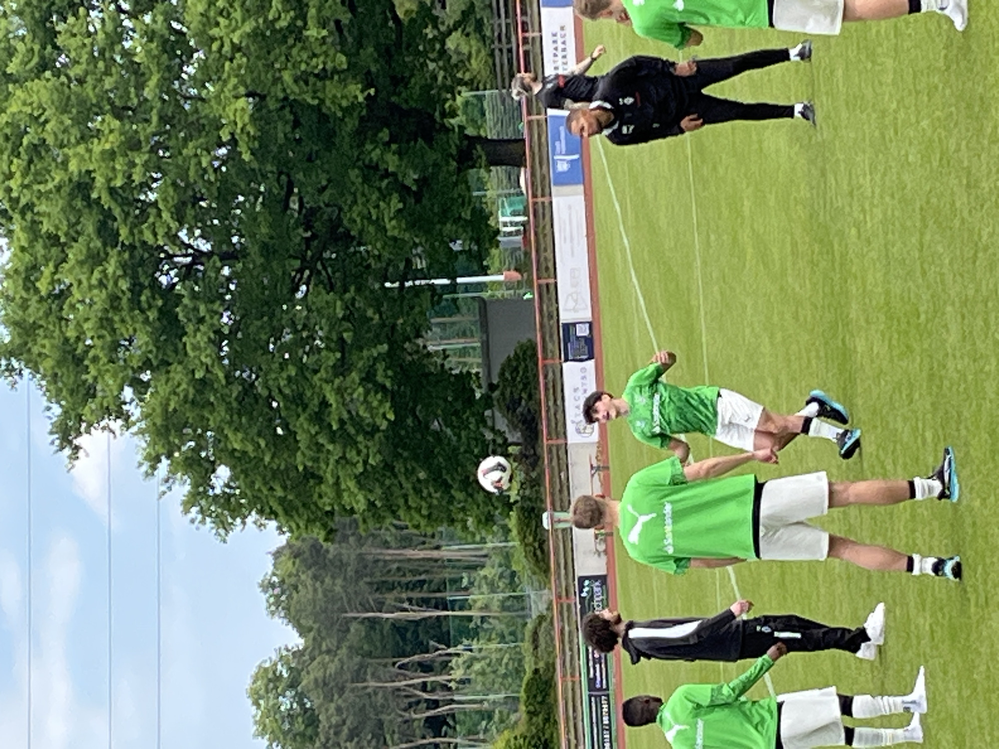 On a soccer field in Kelsterbach, Germany, several soccer players are engaged in training. Players are wearing green shirts with white shorts or black tracksuits. The soccer ball is in the air. Tall trees surround the field and line the fence. The sky is blue.