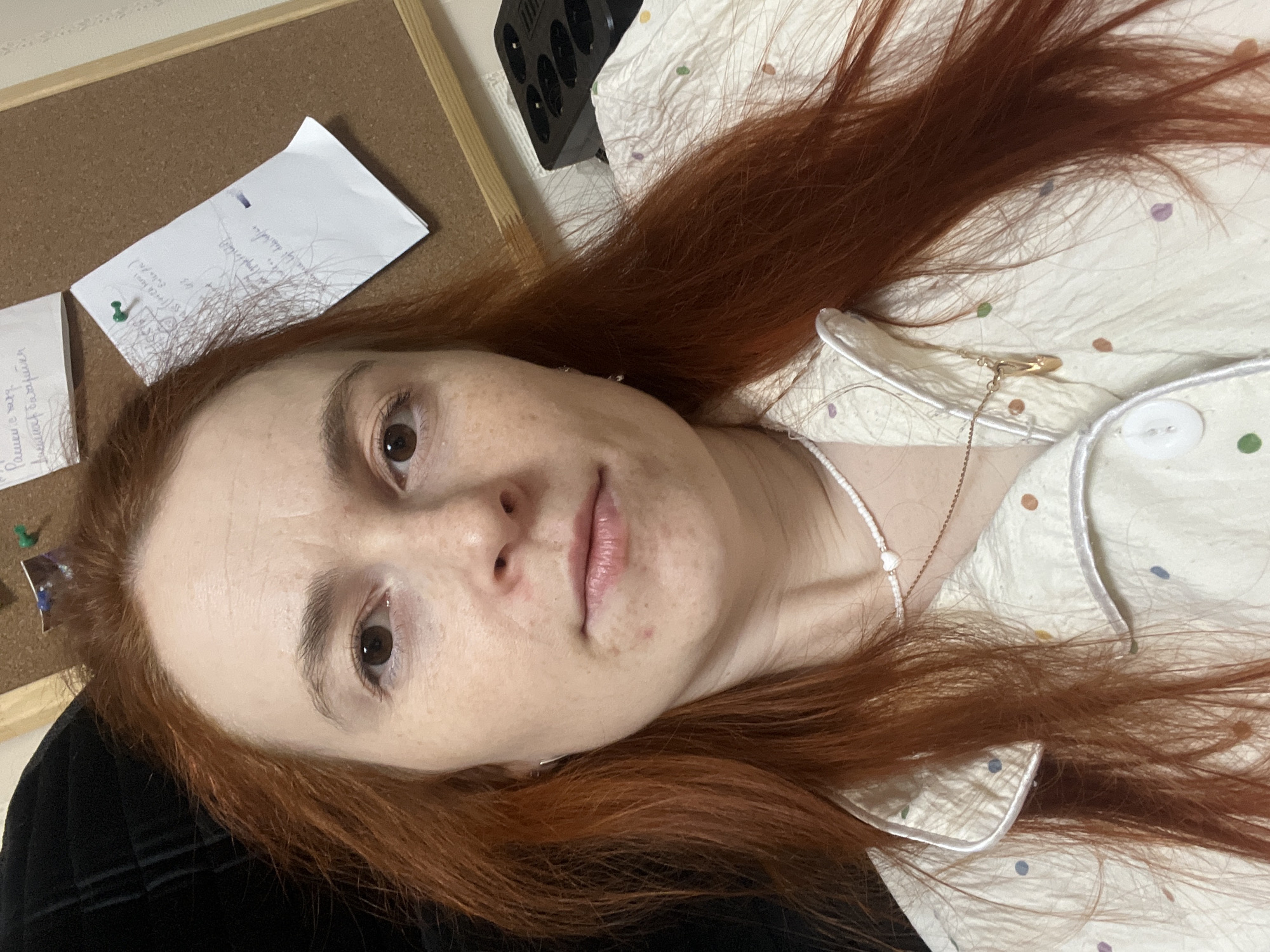 A selfie of a light-skinned woman with red hair. She is lying on a bed wearing a cream-colored pajama top with blue and brown polka dots, a white collar, and white buttons. She is also wearing a white beaded necklace. The background consists of a corkboard with paper attached, a black power strip, and a black garment. There is no verifiable information to indicate the location is Chaikovsky, Russia.