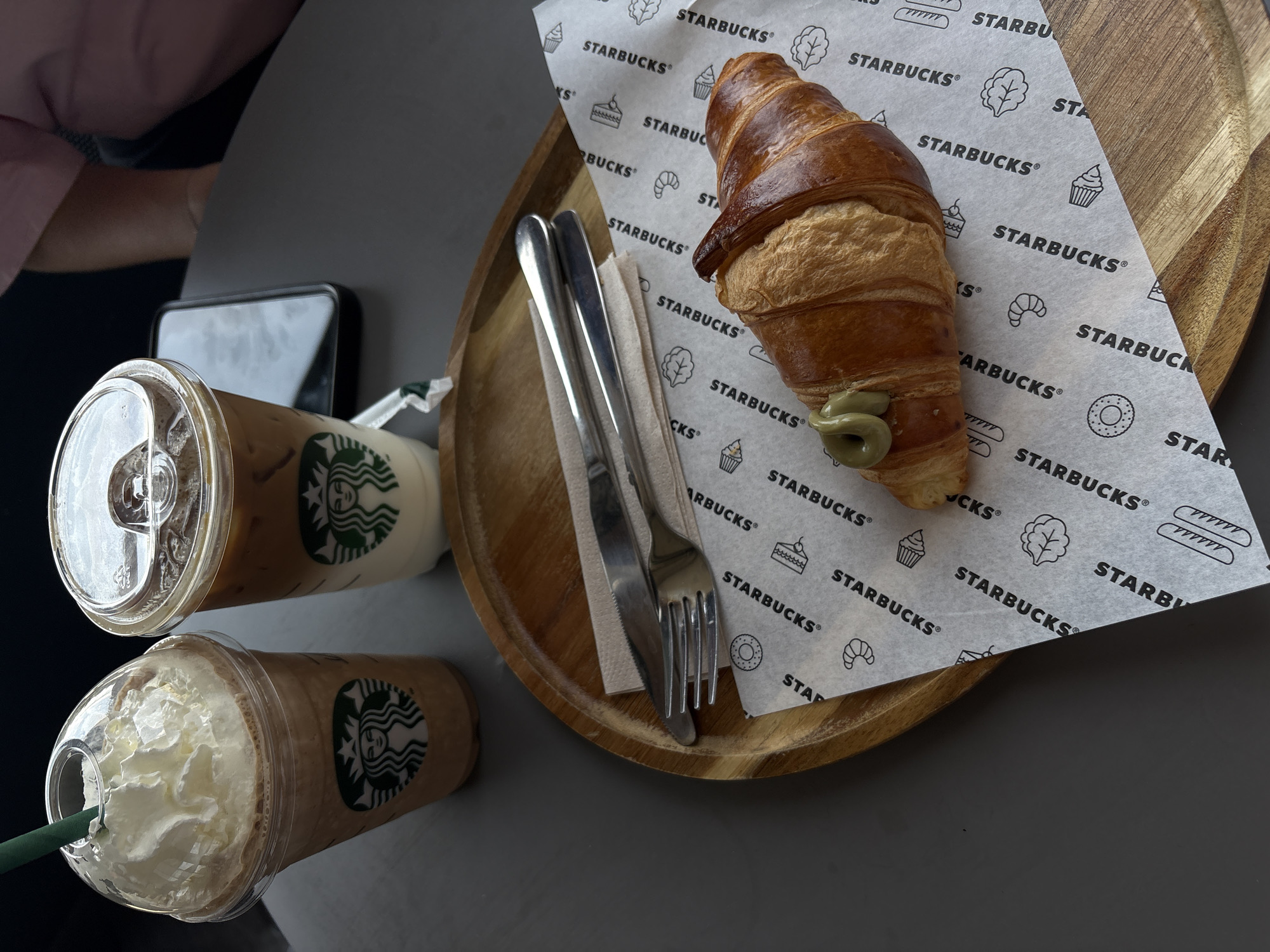 The image displays a casual cafe setting, likely in Plovdiv, Bulgaria, featuring Starbucks beverages and a pastry. Two Starbucks drinks occupy the left side of the frame. The upper drink is an iced coffee in a clear plastic cup with a lid, showcasing its brown liquid. Below it, a blended drink, possibly a Frappuccino, is topped with a generous swirl of whipped cream and has a green straw. Both cups feature the recognizable Starbucks siren logo.
On the right, a golden-brown croissant with a visible green filling or drizzle, possibly pistachio, is presented on a piece of white paper printed with the "STARBUCKS" logo and various line-art icons such as cupcakes, croissants, bread, lettuce, and donuts. This pastry and paper rest on a round, light-colored wooden tray. A set of silverware, consisting of a knife and fork, is placed on a white napkin beside the croissant on the tray.
In the background, a smartphone is partially visible behind the drinks, and a person's arm with a pink sleeve is subtly present in the upper left corner. The table surface is a solid dark gray, contrasting with the warm tones of the wooden tray and croissant. The lighting suggests it is daytime. The visible text predominantly features "STARBUCKS" and "STARBUCKS®" repeatedly on the paper under the croissant.